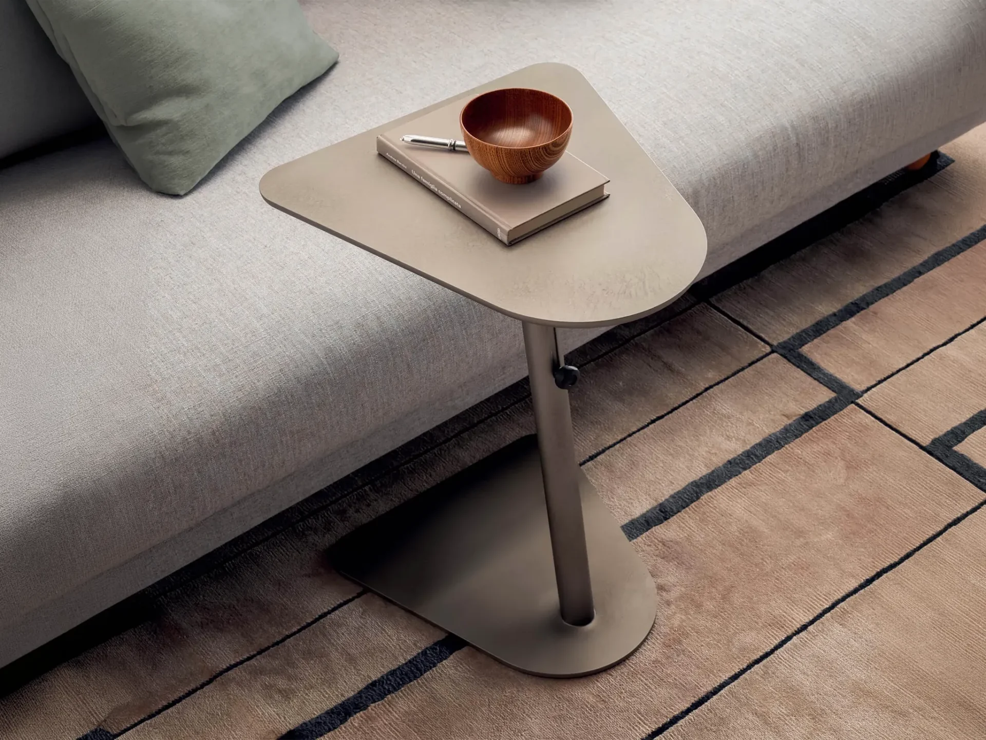 A triangular side table next to a light gray sofa. On the table are a book, a silver pen, and a wooden bowl. A light brown rug with dark geometric lines covers the floor.
