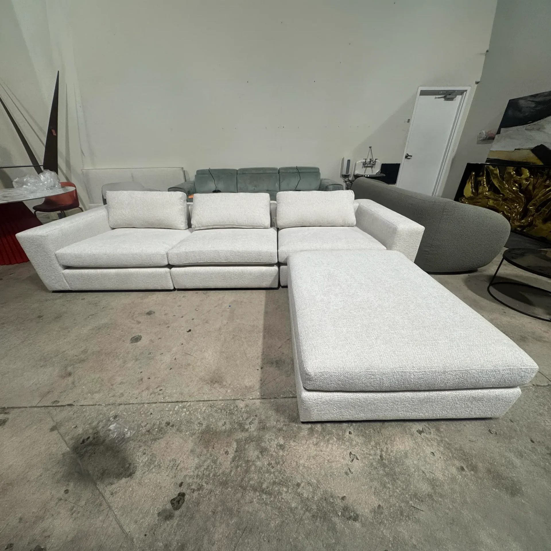 A light grey modular sectional sofa with an ottoman on a concrete floor, surrounded by other furniture in a showroom.