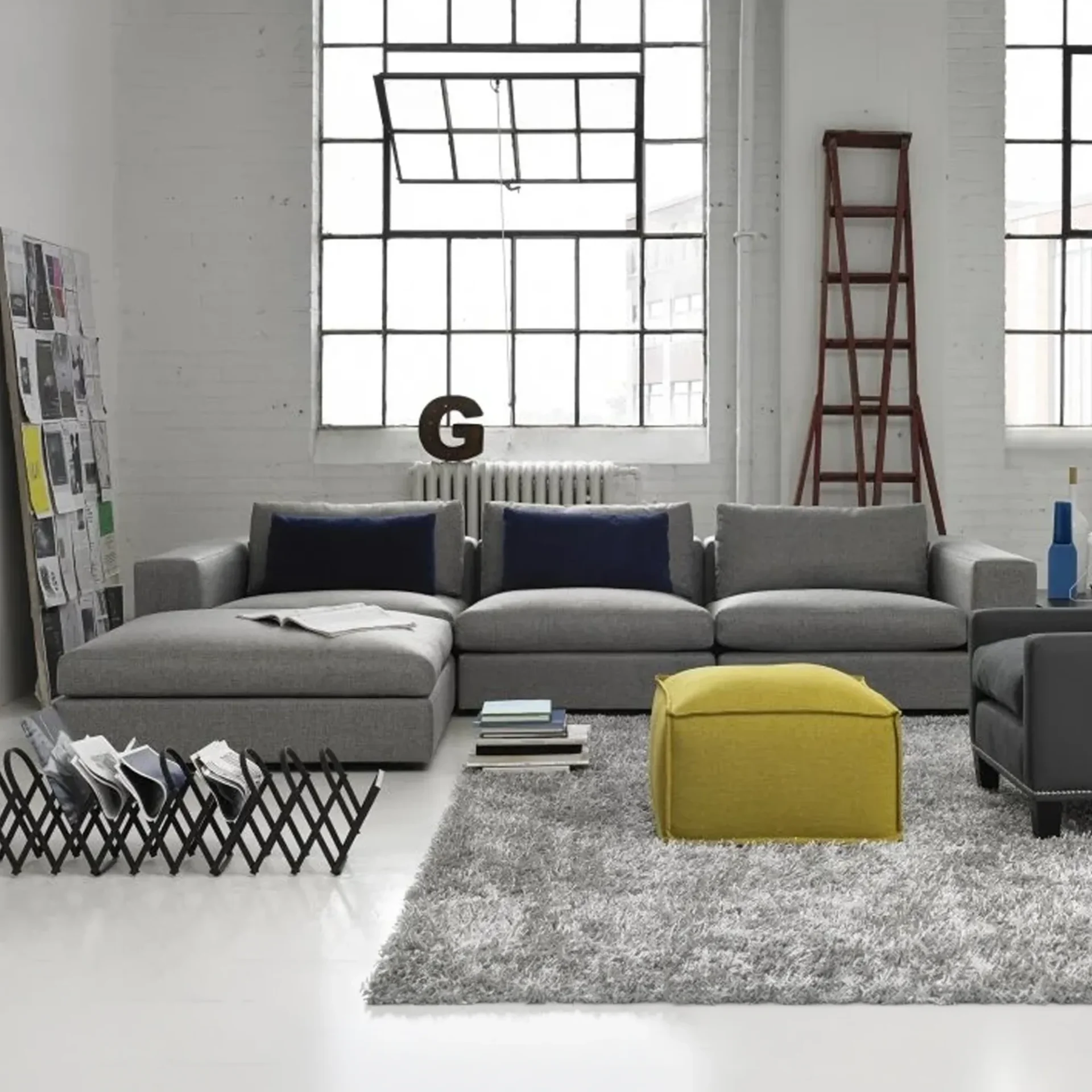 A modern living room with a grey L-shaped sectional sofa, a yellow ottoman, a grey shag rug, and large industrial windows.
