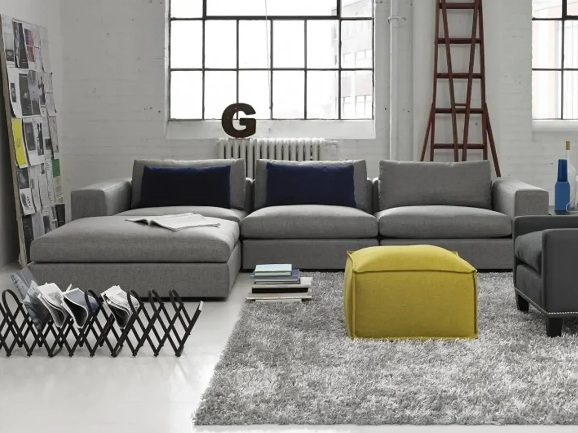 A modern industrial living room with a gray sectional sofa, a yellow ottoman, a gray shag rug, and large black-framed windows.