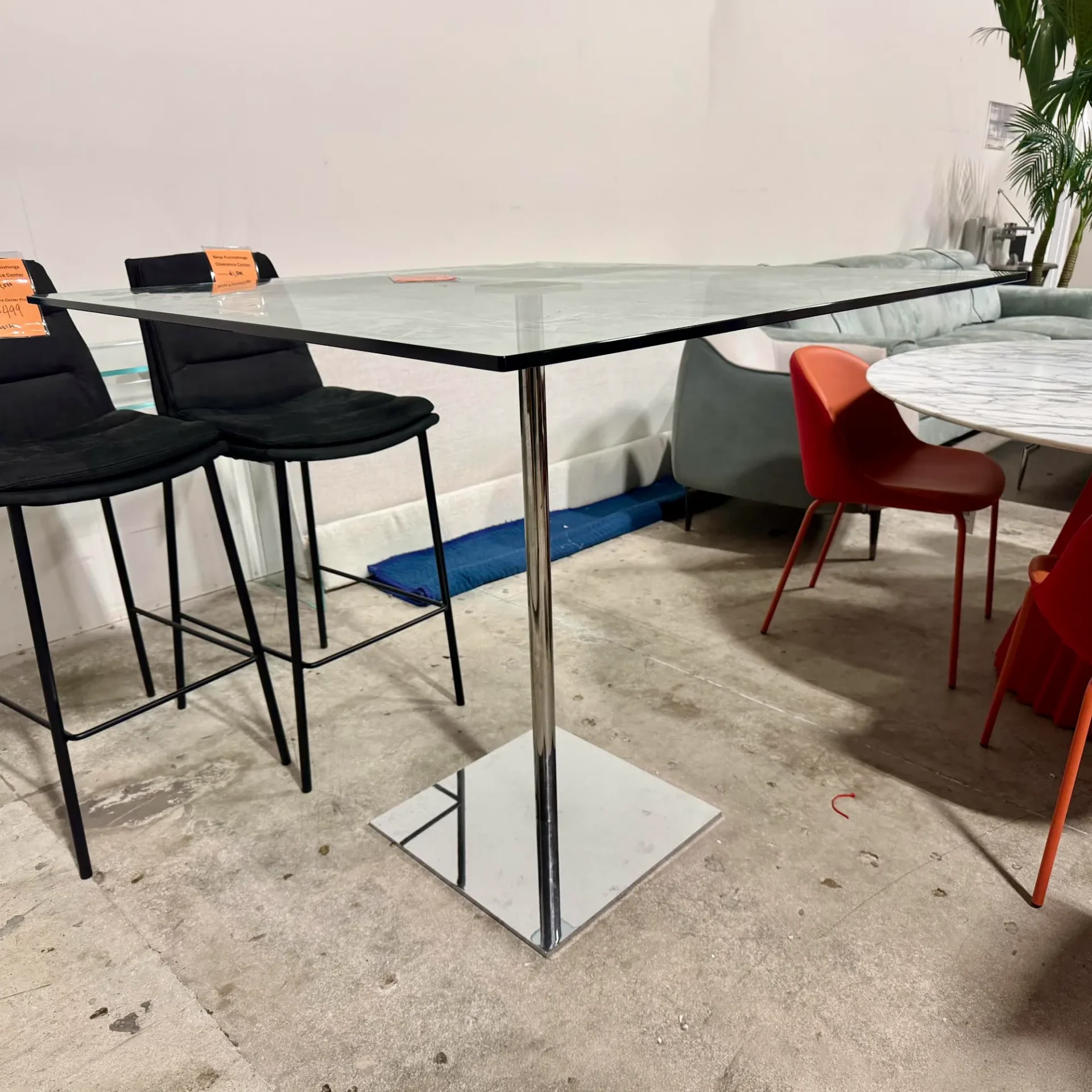 A square glass-top bar table with a chrome base, flanked by two black bar stools, in a showroom setting.