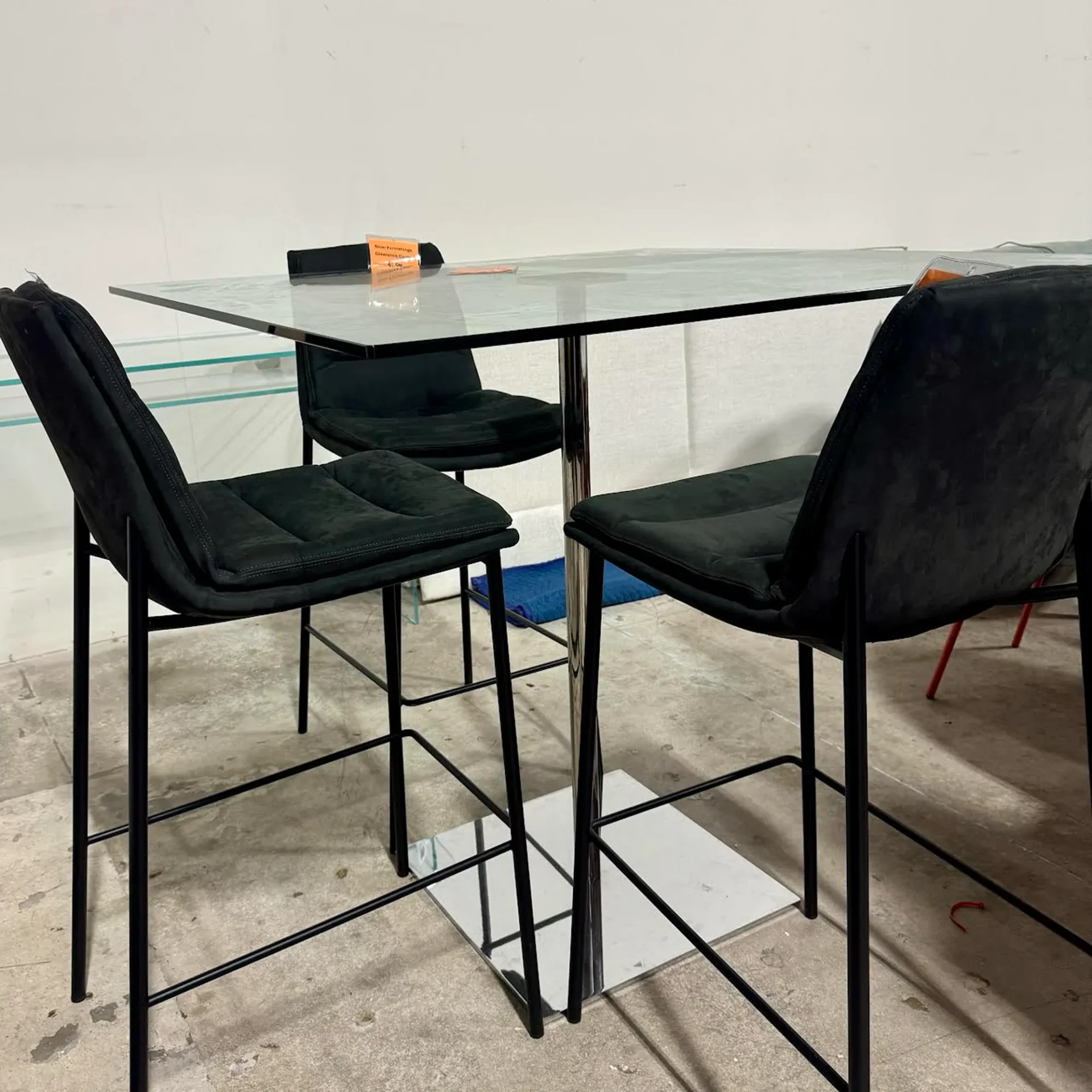 A rectangular glass bar table with a chrome pedestal base, surrounded by three black upholstered bar stools with black metal frames.