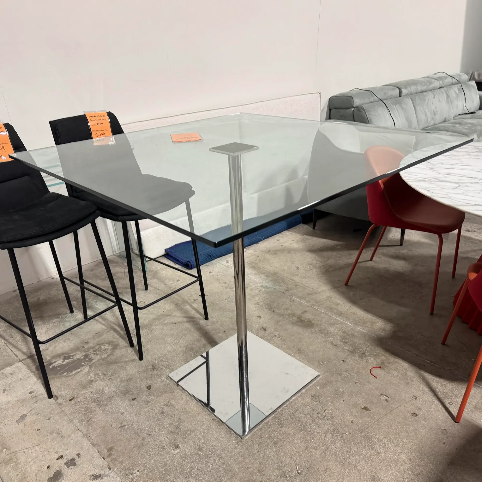 A square glass-top table with a chrome pedestal base, two black bar stools, a gray sofa, and red chairs on a concrete floor in a furniture showroom.