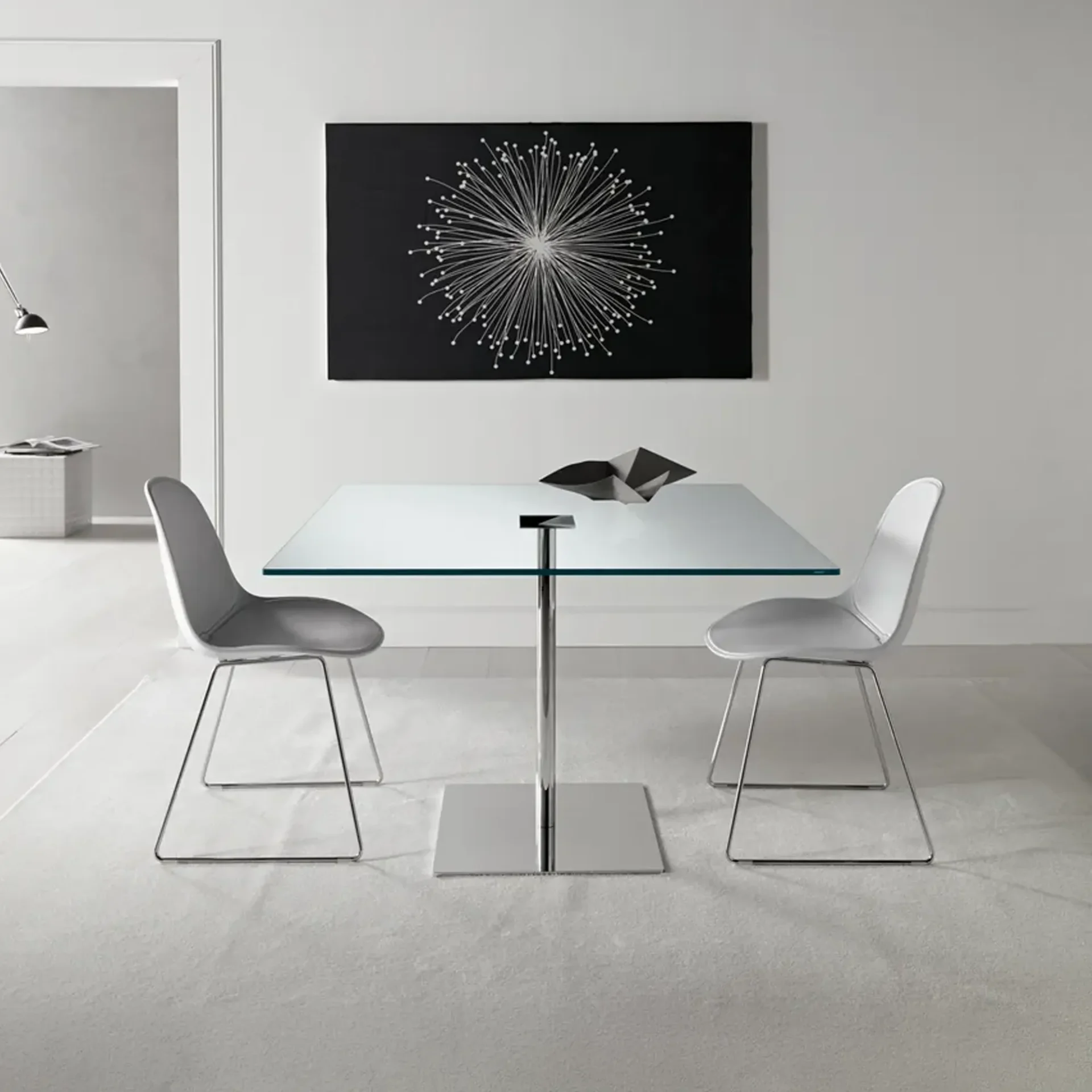 A modern dining room with a square glass table, two light gray chairs, and a black and white starburst artwork on the wall.