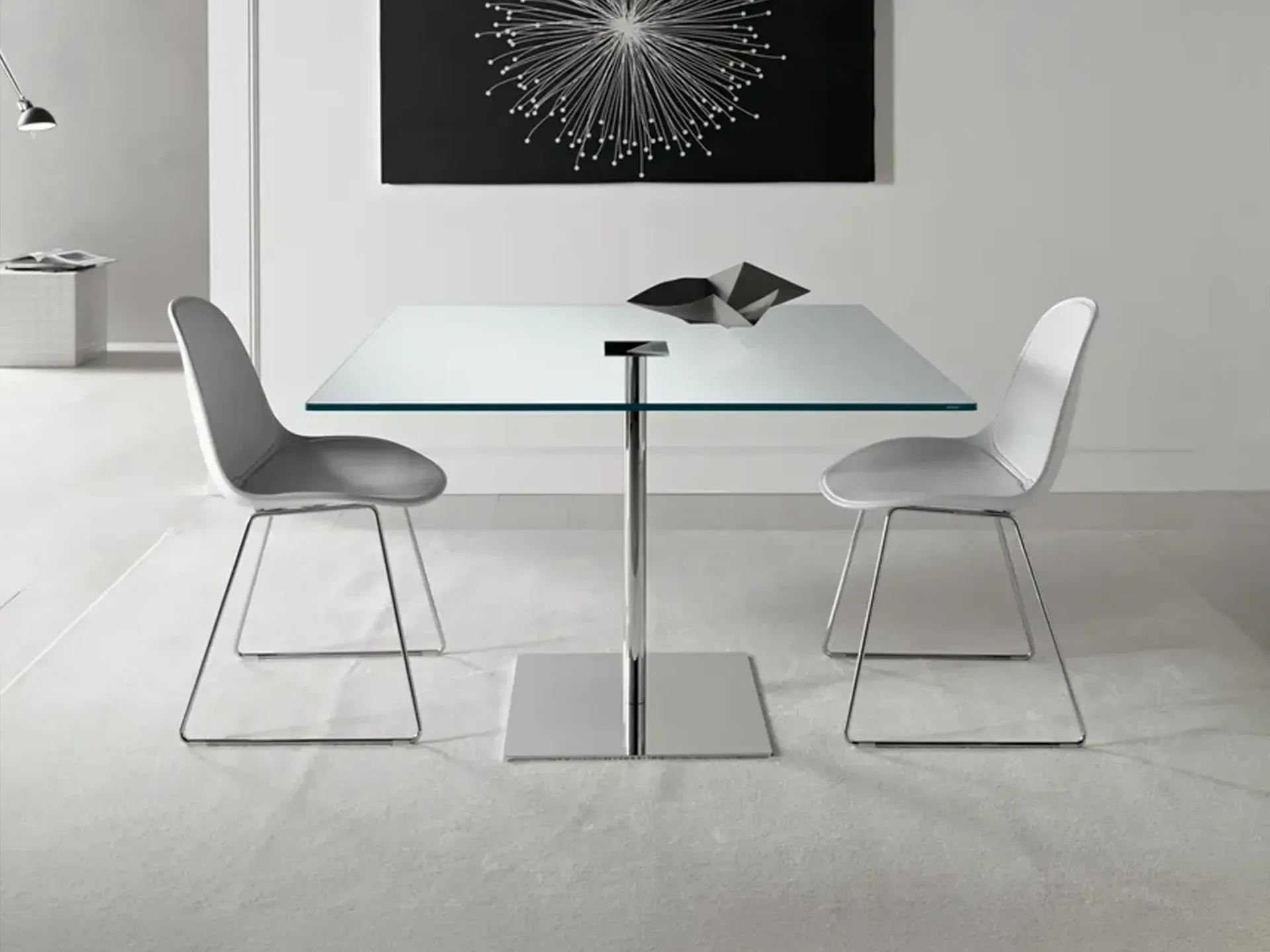 A modern dining setup with a square glass table, two gray chairs, and a black abstract artwork on a white wall.