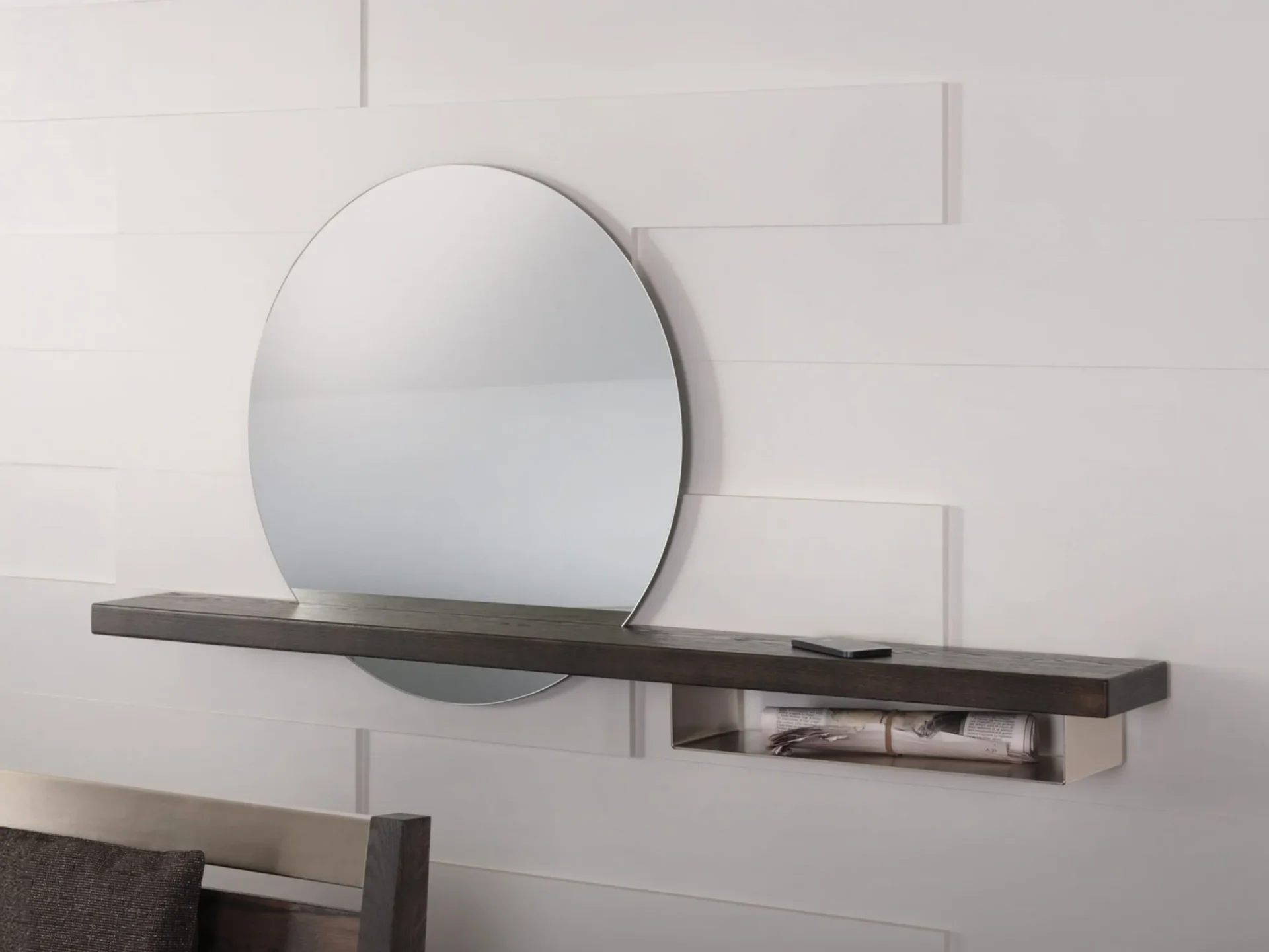 A large oval mirror above a dark wood floating shelf with a hidden compartment holding rolled newspapers, against a white paneled wall.