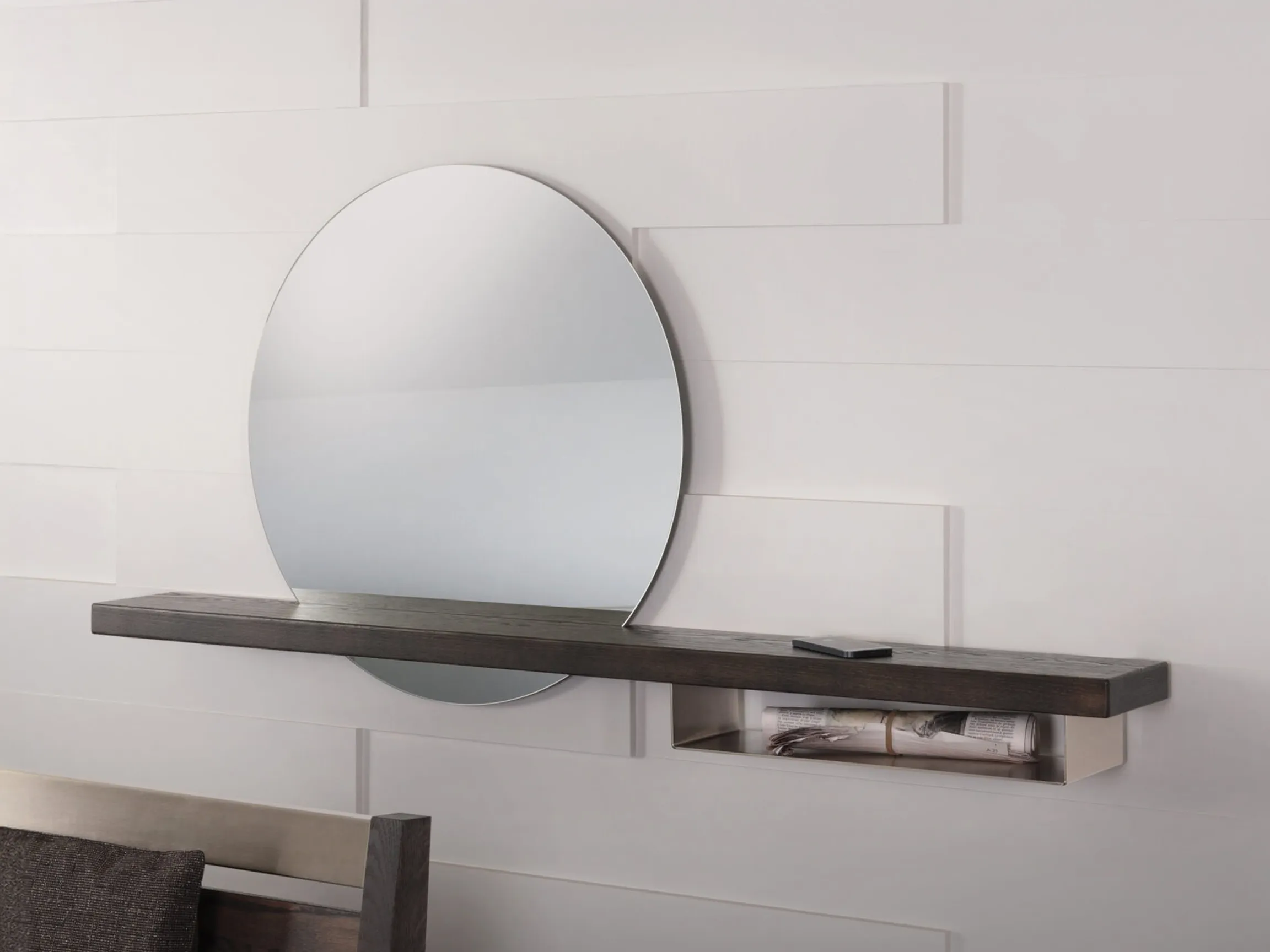 A large oval mirror above a dark wood floating shelf with a hidden compartment holding rolled newspapers, against a white paneled wall.
