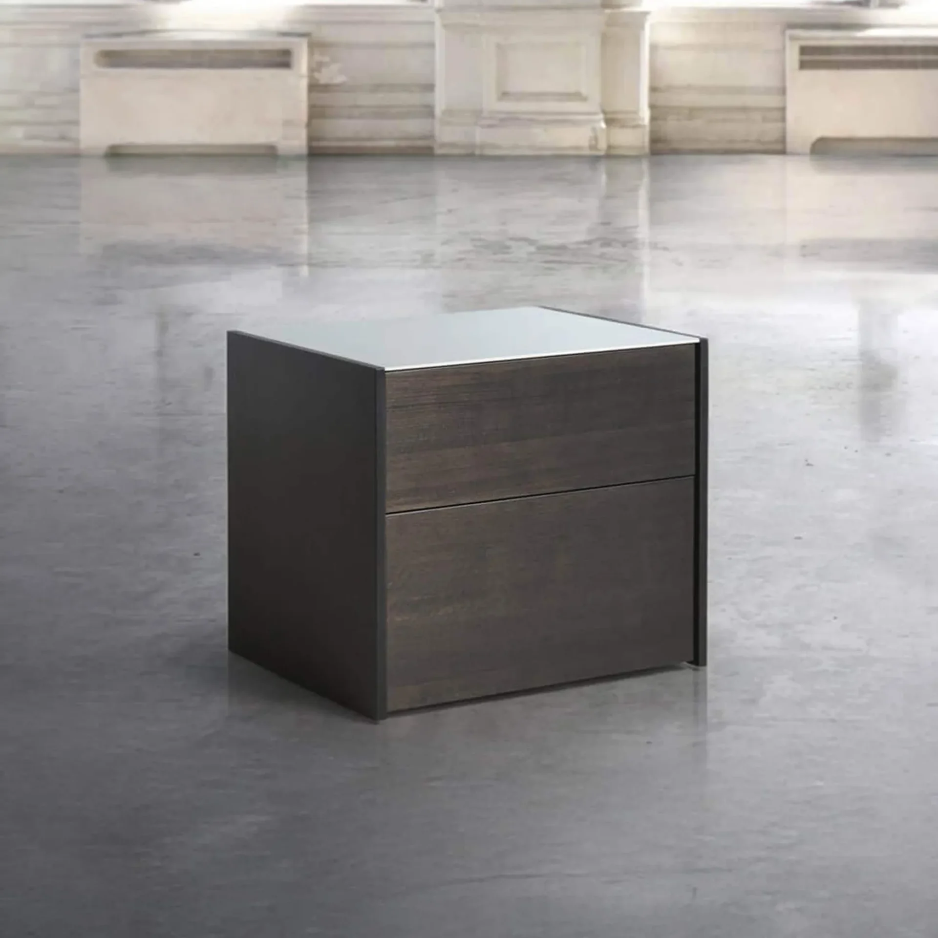 A dark brown two-drawer cabinet with a reflective light-colored top, standing on a polished gray floor in a room with light architectural walls.
