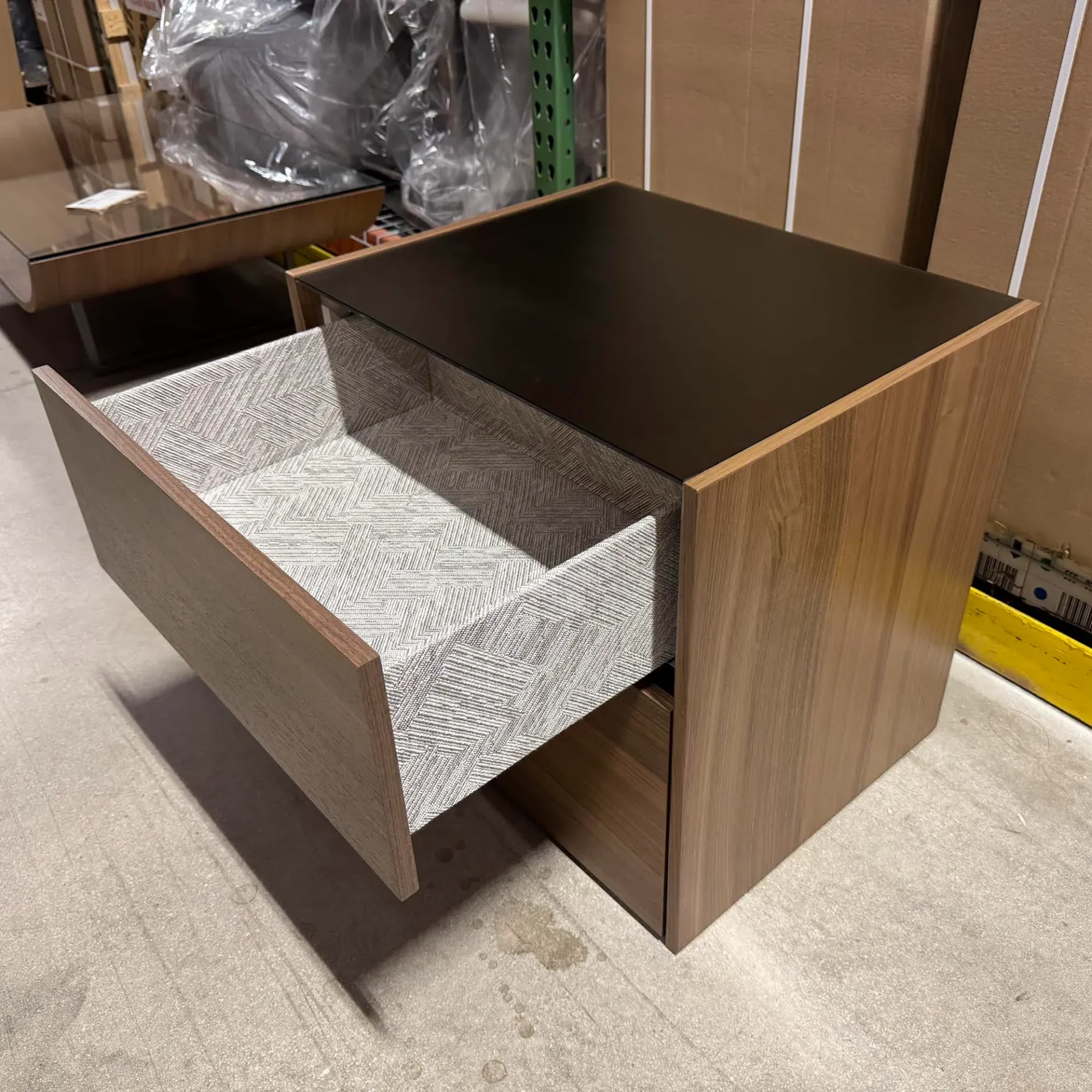 A modern, light wood nightstand with a dark top and an open top drawer revealing a patterned interior. It sits on a gray floor in what appears to be a warehouse setting.