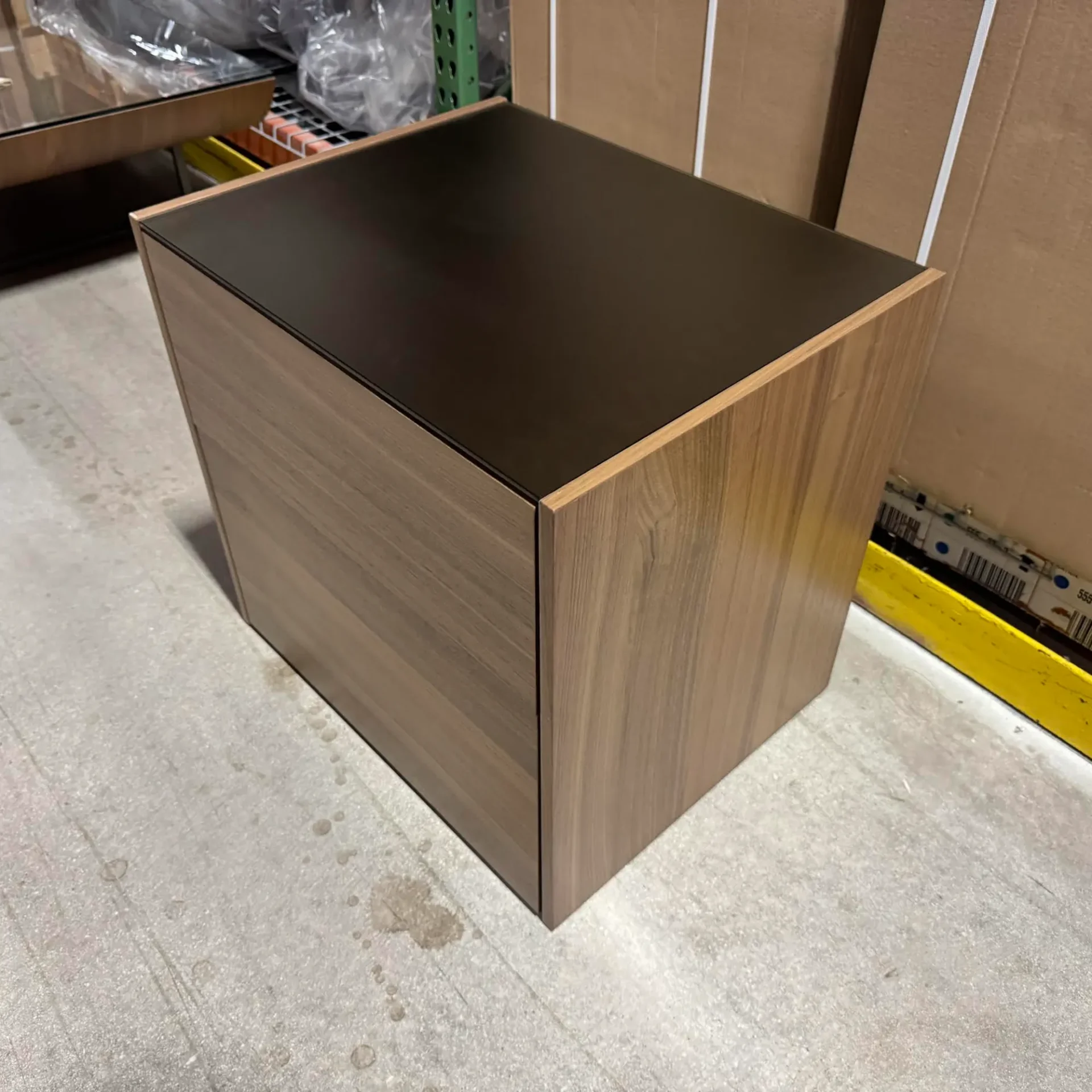 A square side table with a dark, reflective top and light wood-grain sides, placed on a concrete floor in an industrial setting.