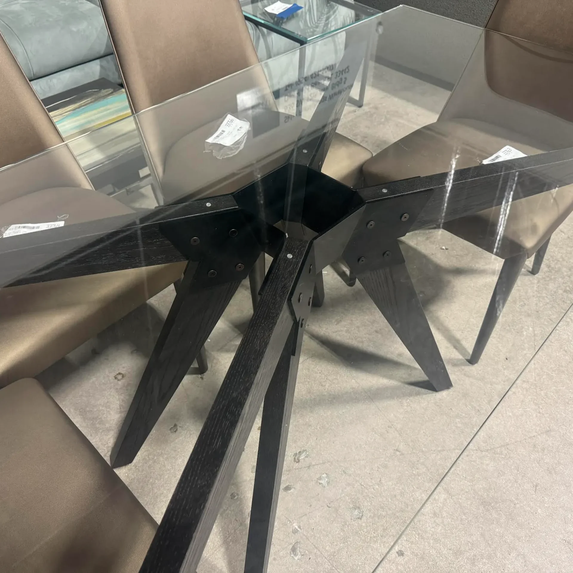 A modern dining table with a clear glass top and a dark, geometric wooden base, surrounded by taupe upholstered chairs on a light gray floor.