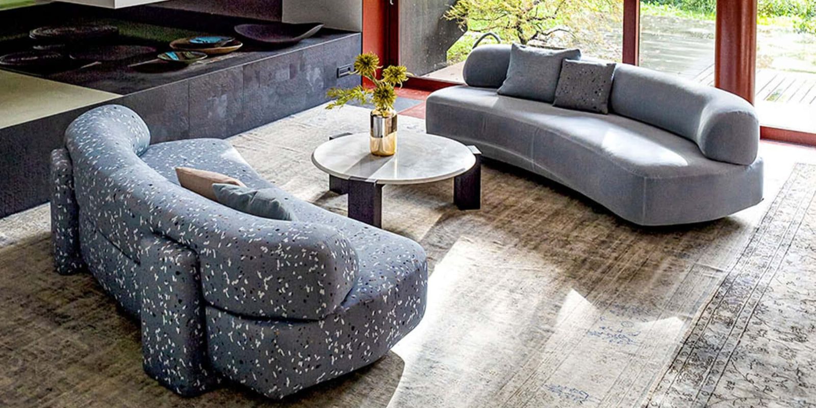 A modern living room with two curved sofas, a round coffee table with a vase of foliage, and a patterned rug, illuminated by natural light from large windows overlooking a garden. Gogan Modular Curved Sofa