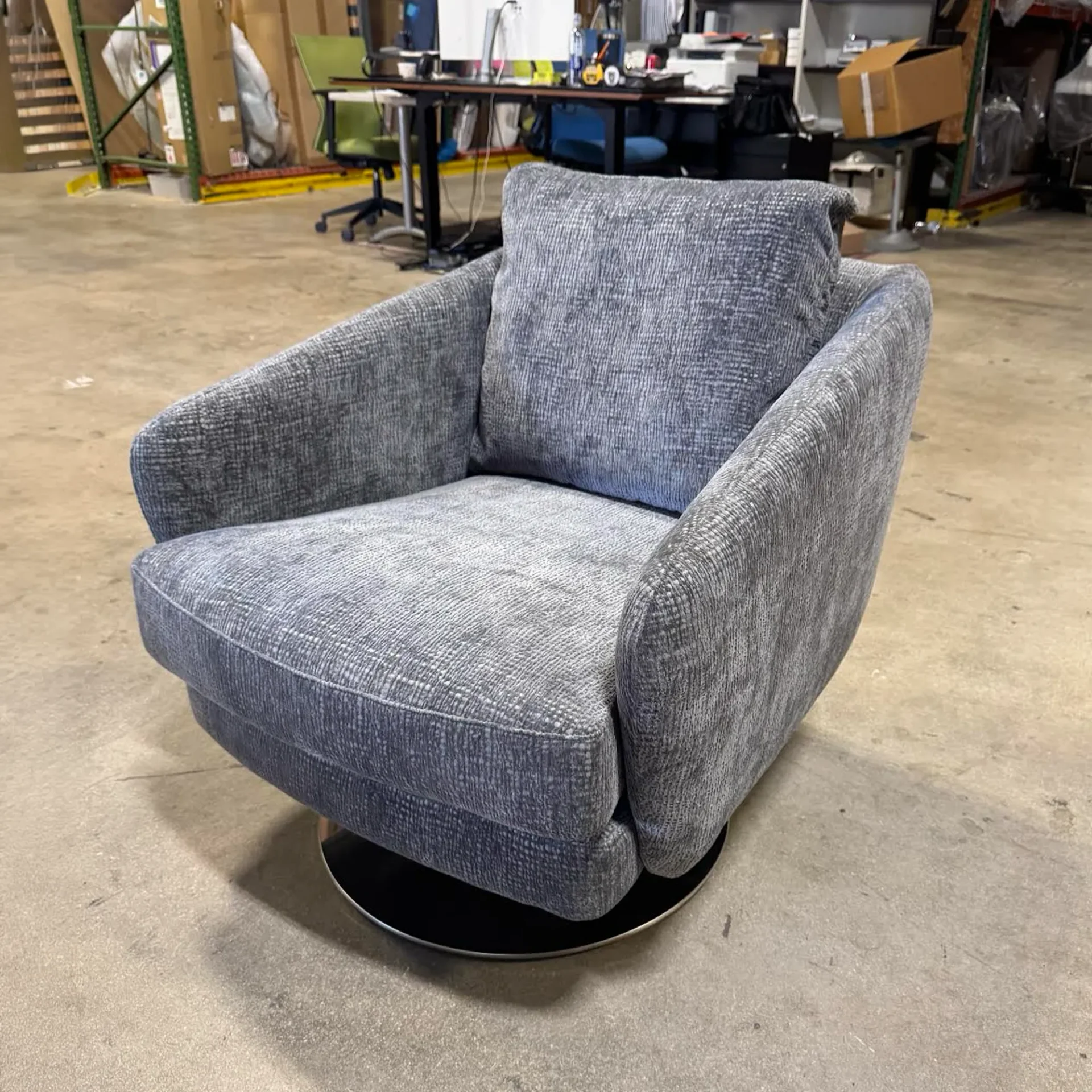A gray upholstered swivel armchair with a textured fabric and a black circular base on a concrete floor.