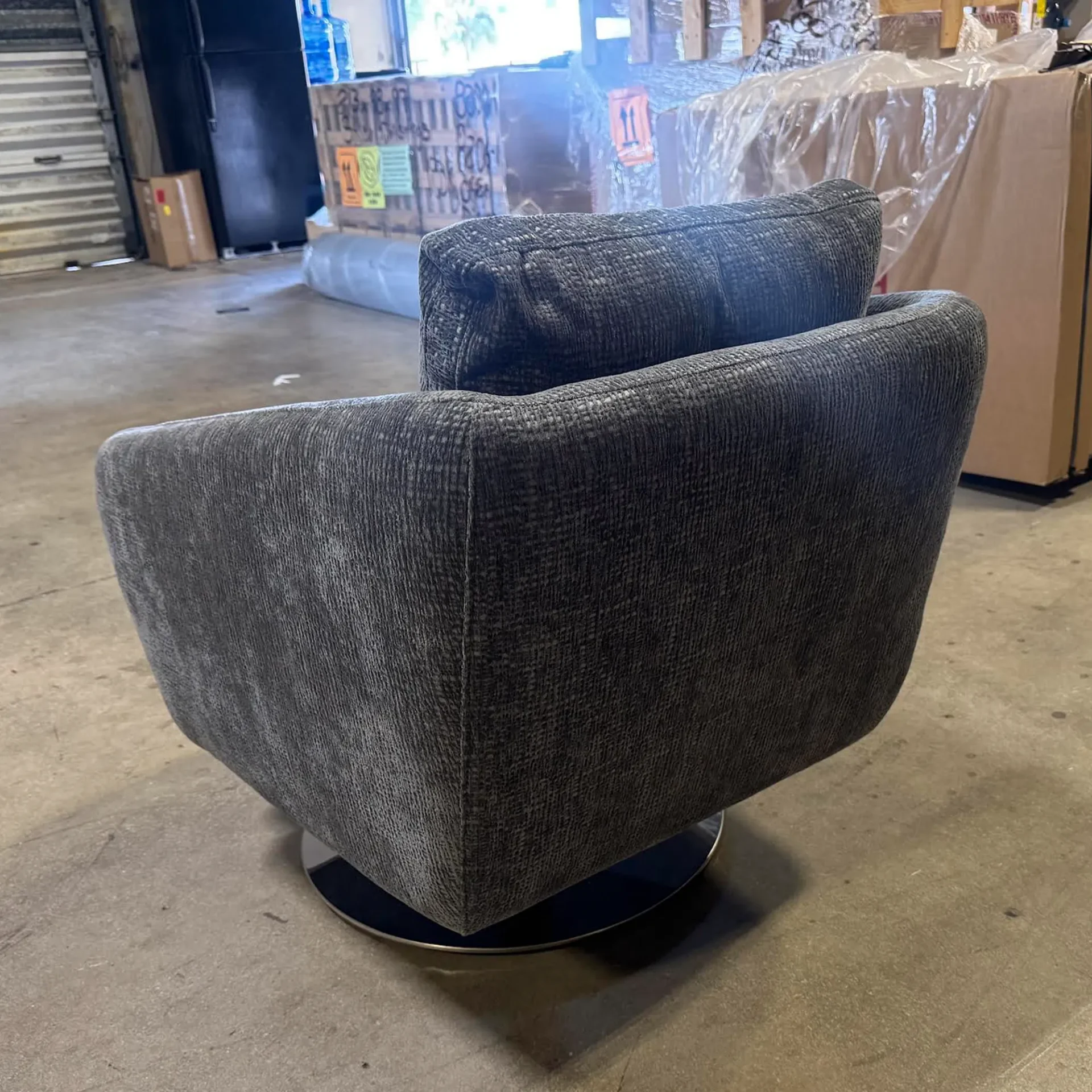 A dark gray textured fabric swivel chair with a rounded back and a circular base, sitting on a concrete floor in a warehouse setting.