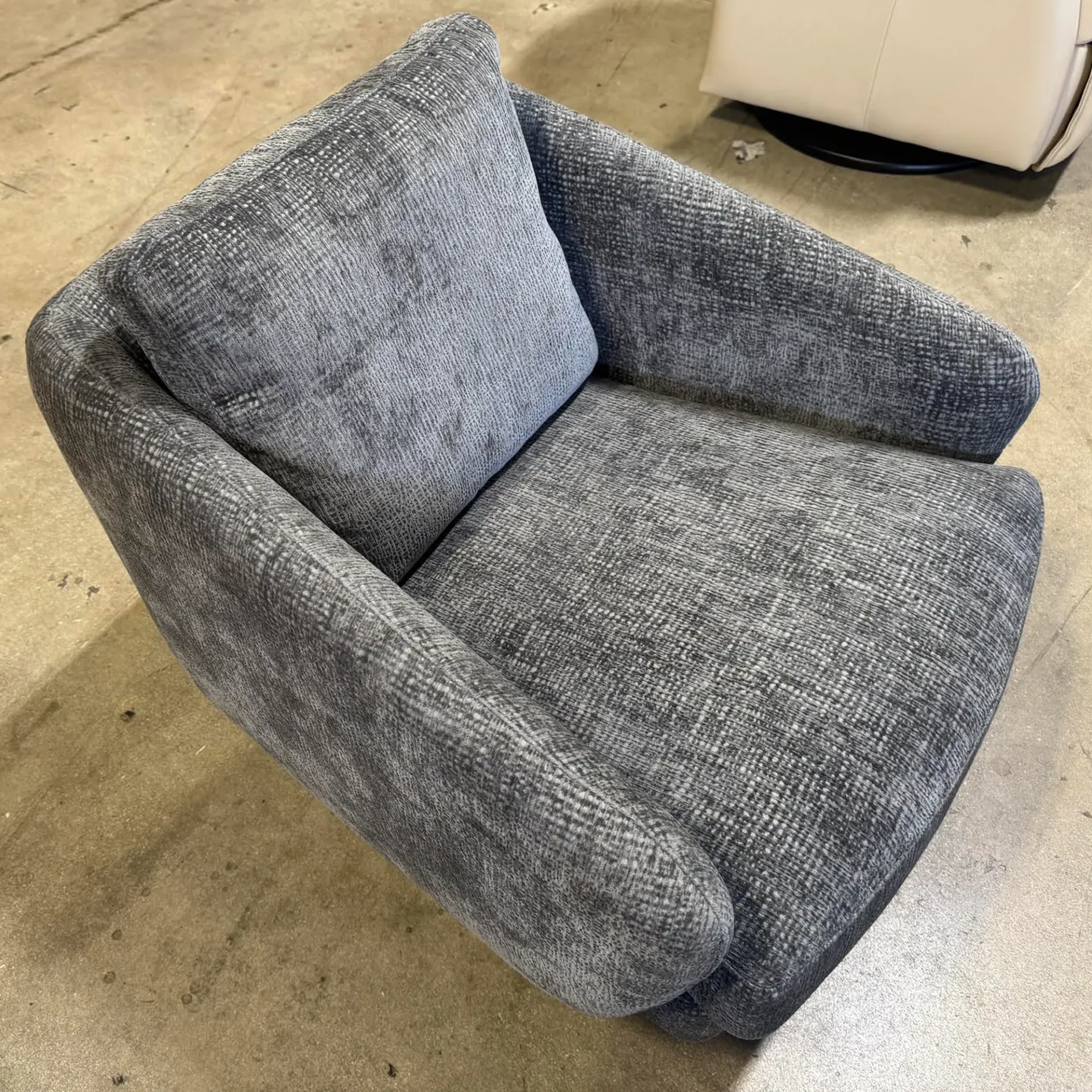 A gray, textured, rounded armchair with a separate back cushion sits on a concrete floor.