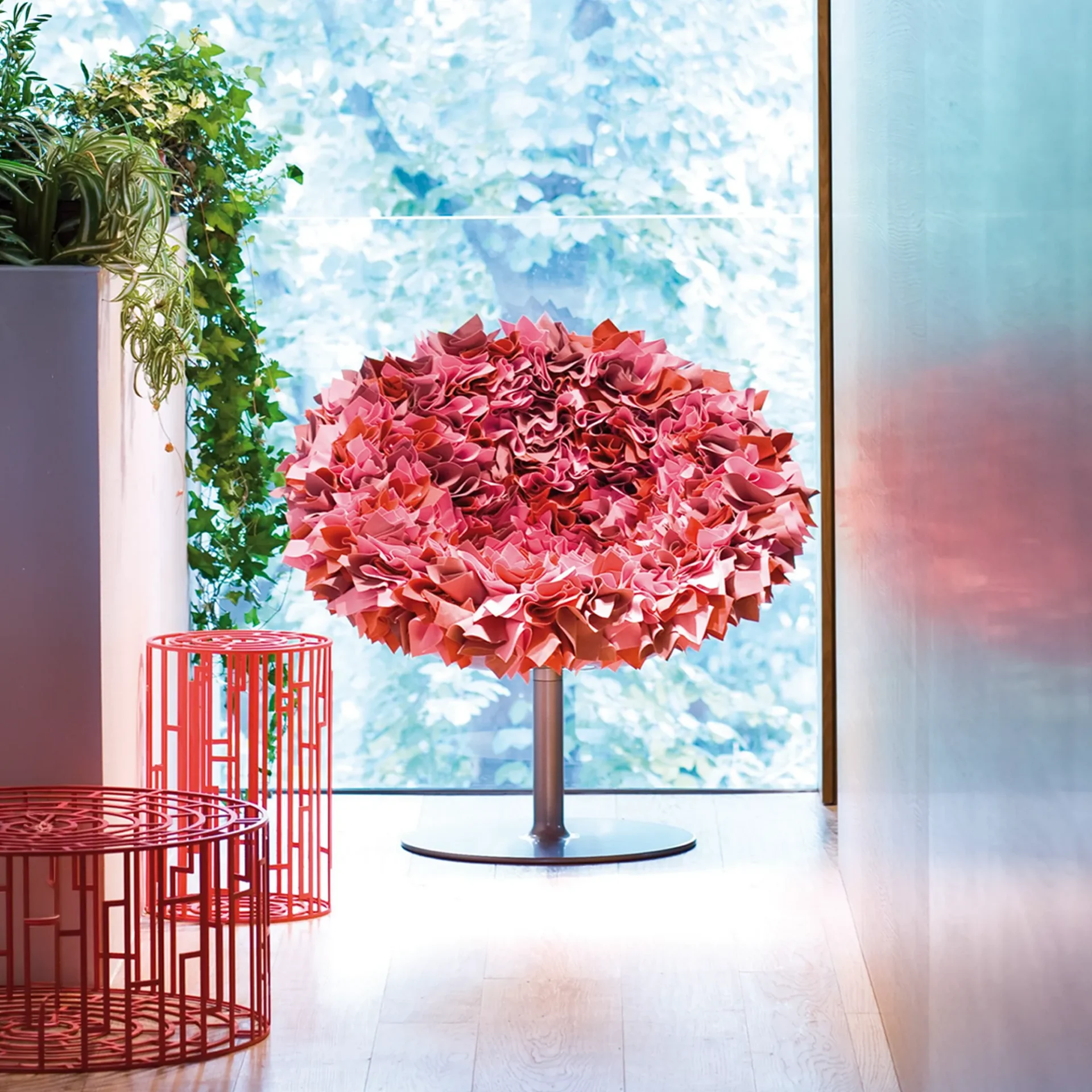 A large, round chair covered in ruffled pink fabric sits in front of a window with blurred green foliage. Two red geometric tables and potted plants are to the left.