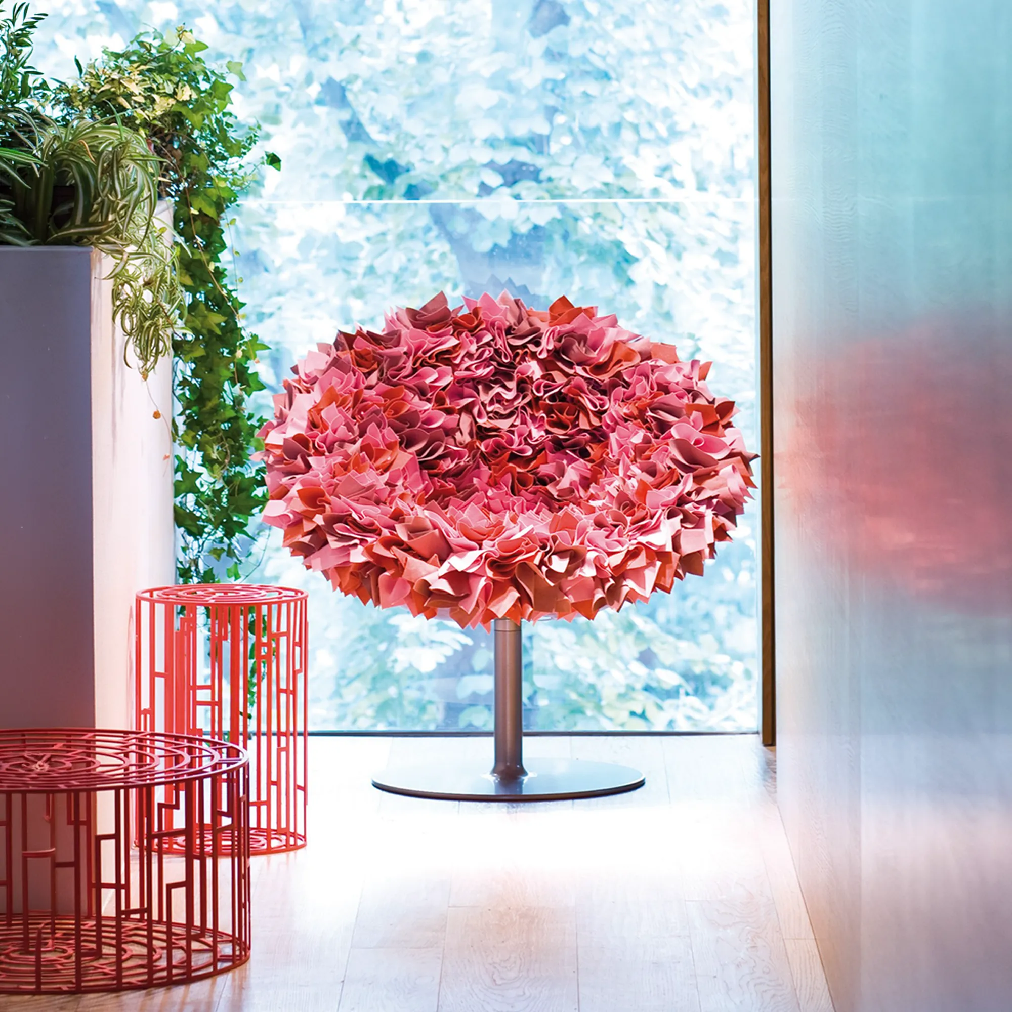 A large, round chair covered in ruffled pink fabric sits in front of a window with blurred green foliage. Two red geometric tables and potted plants are to the left.