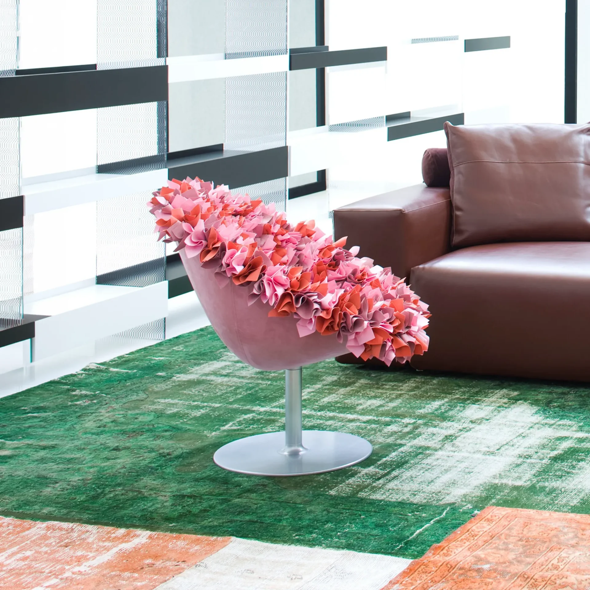A pink ruffled swivel chair on a green and orange rug, with a brown leather sofa and geometric room divider in the background.