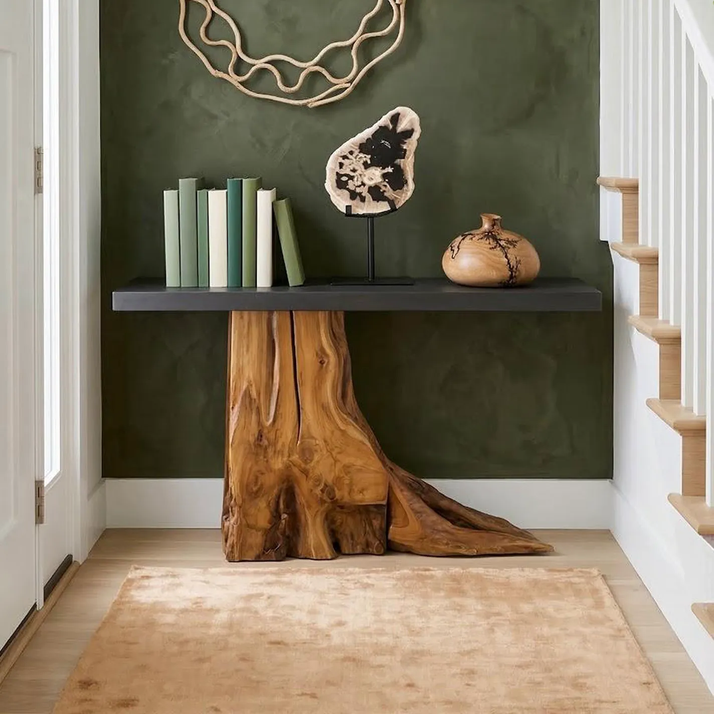 An entryway featuring a console table with a dark gray top and a natural live-edge wood base against a dark green wall. The table holds books, a petrified wood slice, and a wooden vase. A light beige rug covers the hardwood floor, and a white staircase is visible on the right.