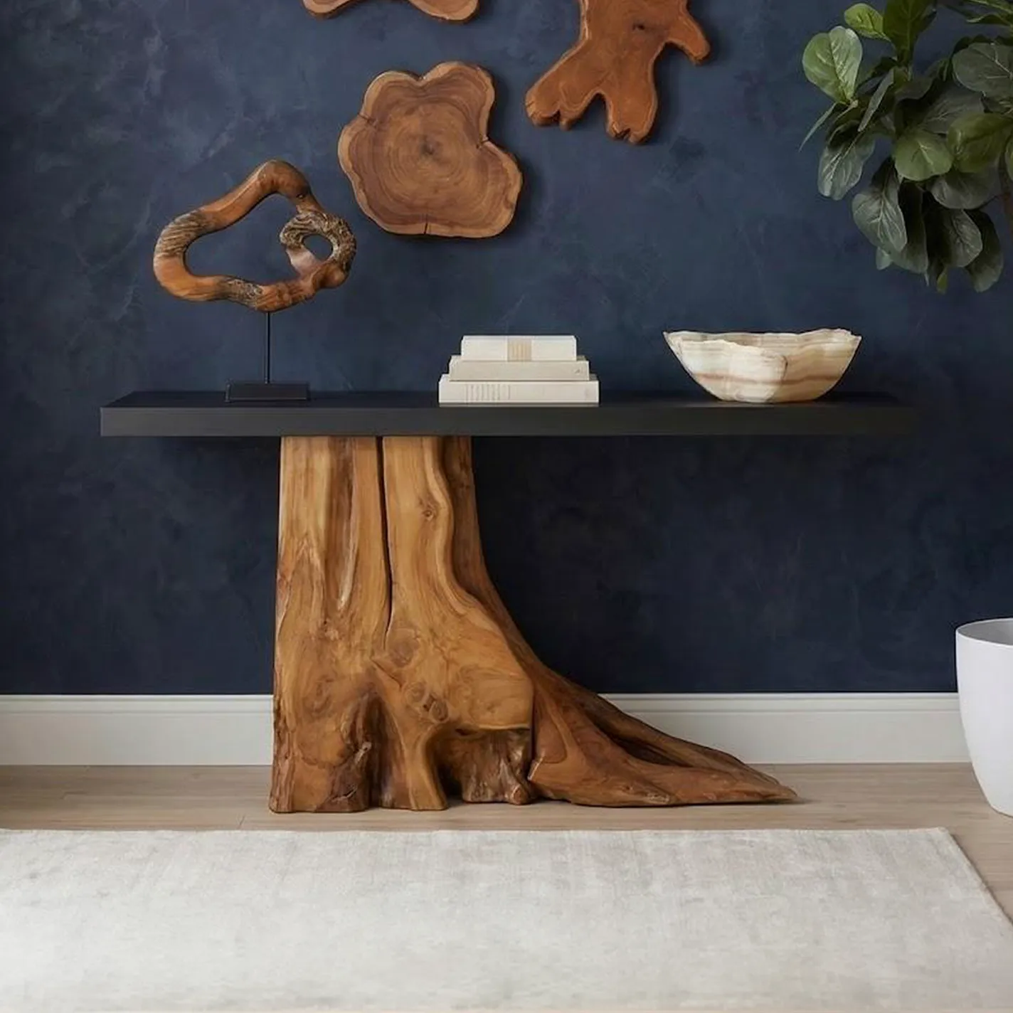 A console table with a natural wood root base and dark top, decorated with a wooden sculpture, books, and a stone bowl, against a dark blue wall with wood art.