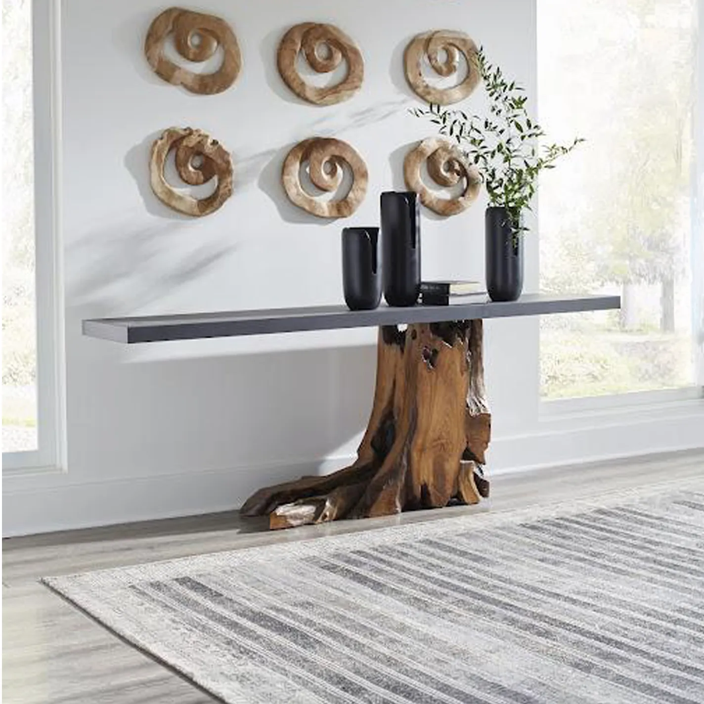 A console table with a dark gray top and a natural, gnarled wood base, adorned with black vases and green foliage, sits against a white wall with six spiral wooden art pieces. A striped gray rug is on the light wood floor.