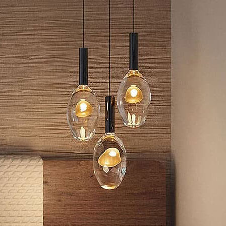 Three modern pendant lights with clear oval glass shades and golden glowing interiors hang against a wooden wall.