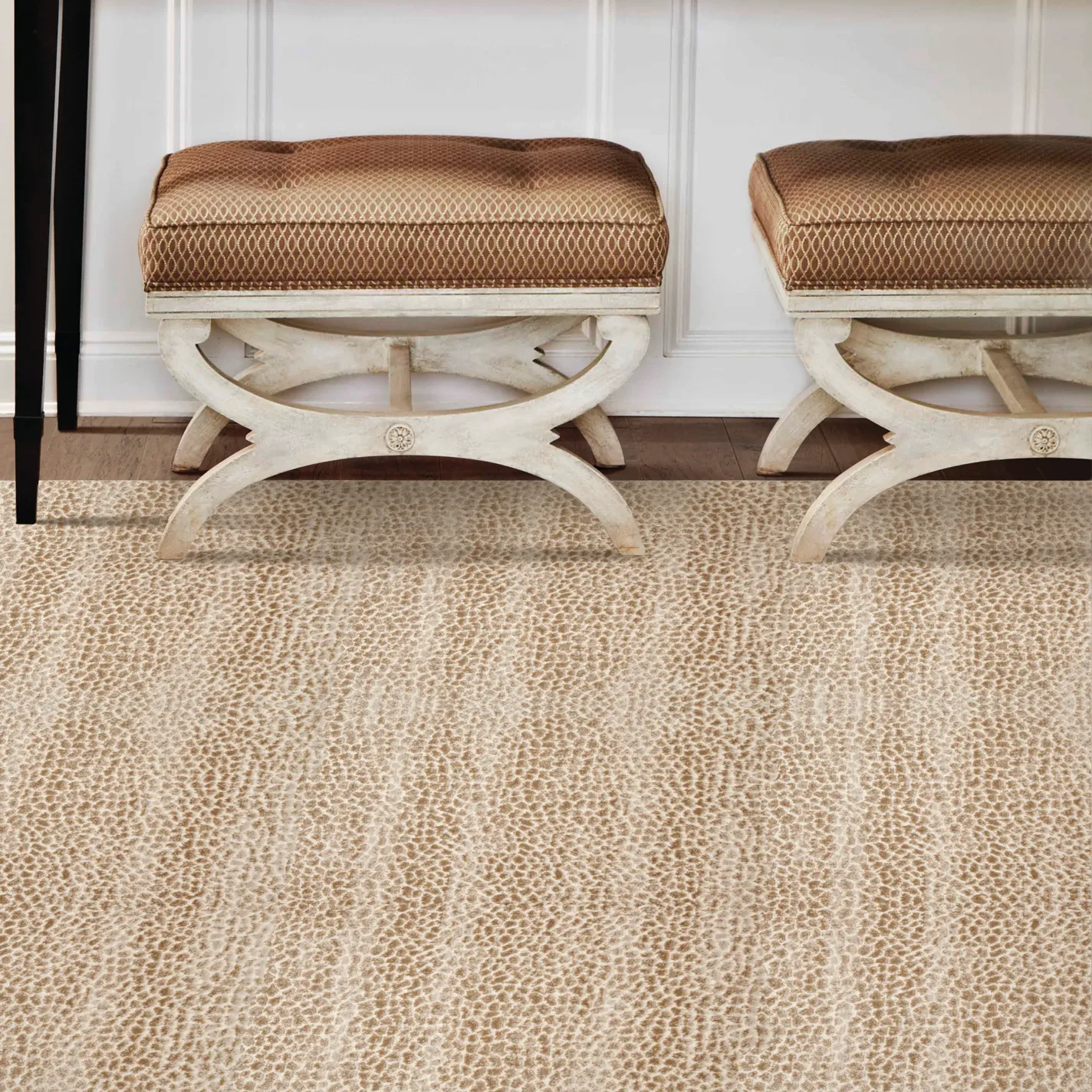 Two X-bench ottomans with patterned brown cushions and distressed white frames sit on a light beige, mottled carpet in front of a white paneled wall. A dark furniture leg is visible on the left.