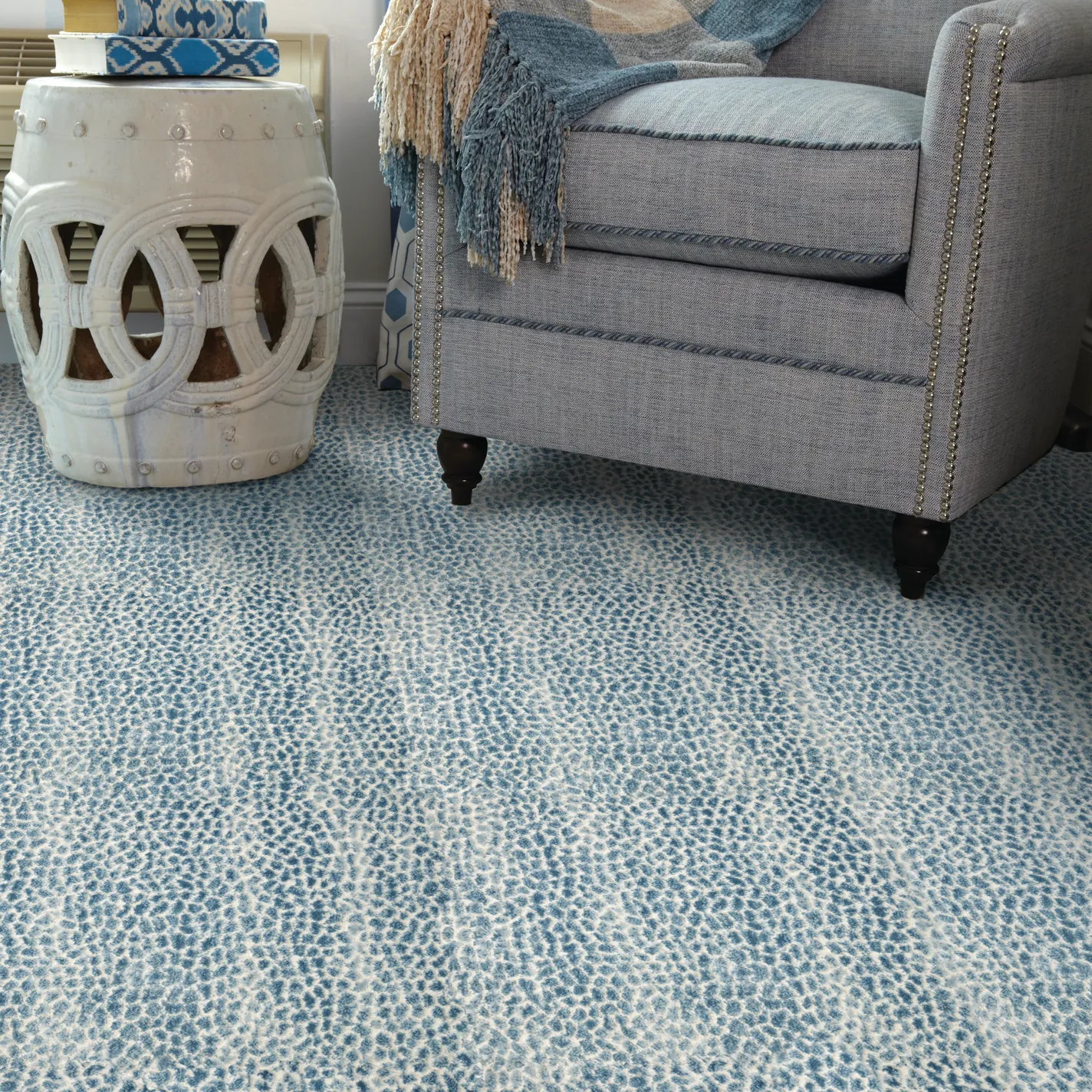 A blue and white patterned carpet with an organic, mottled design. A gray upholstered armchair with nailhead trim and a blue throw blanket is on the right, and a white ceramic garden stool with books is on the left.