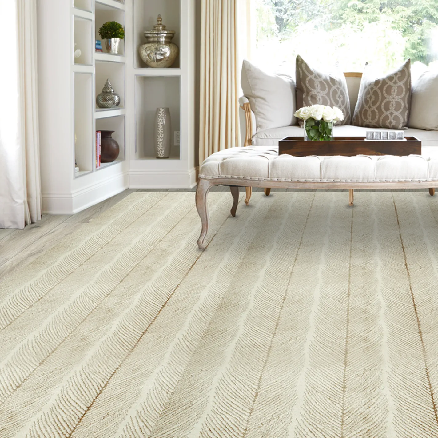 A bright living room with cream-colored textured flooring, a tufted ottoman with white roses, a sofa with patterned pillows, and built-in shelves with decor.
