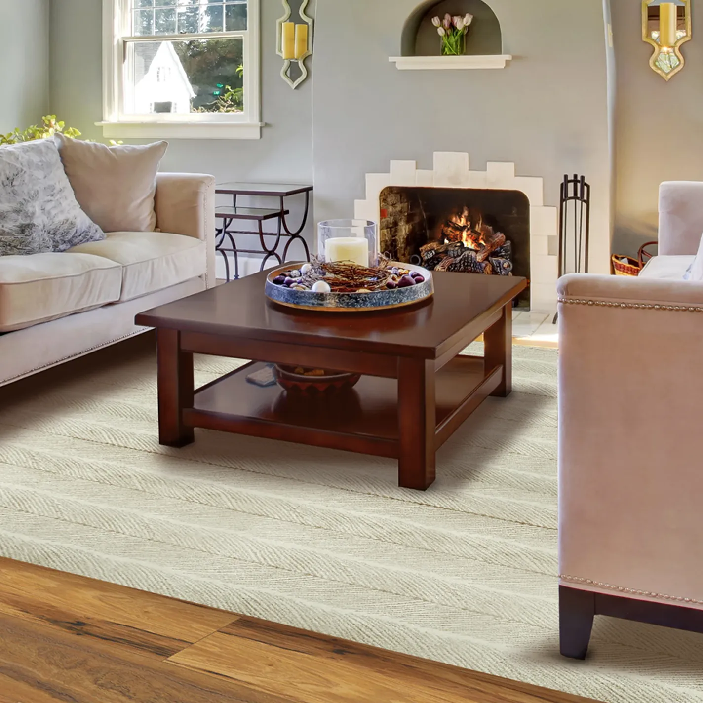 A cozy living room with a cream sofa, dark wood coffee table, and a lit fireplace. A light cream chevron rug covers hardwood floors.