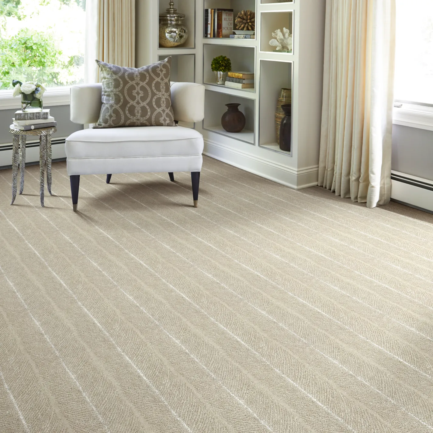 A bright room with light beige diagonal herringbone carpet, a white armchair with a patterned pillow, a silver side table, and built-in white shelves with decor.