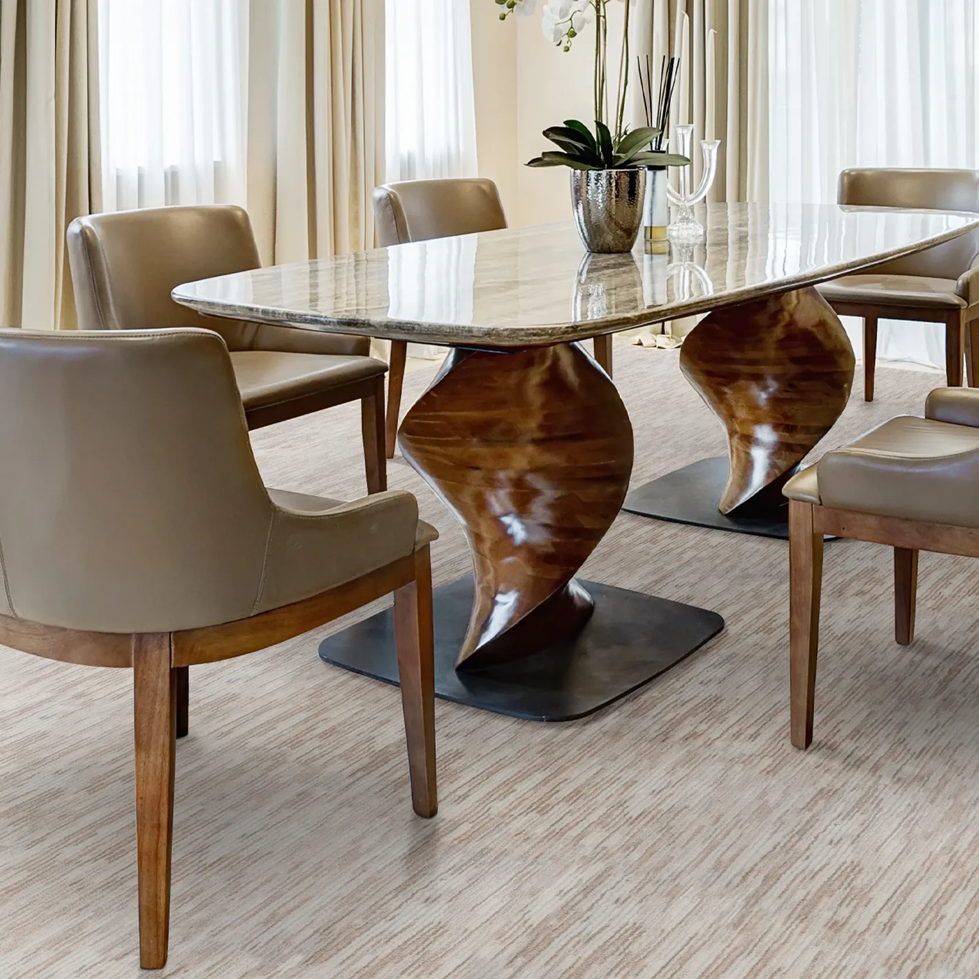 An elegant dining room with a marble-top table on sculptural wooden bases, surrounded by taupe leather dining chairs. The room has light-colored carpet and sheer curtains.