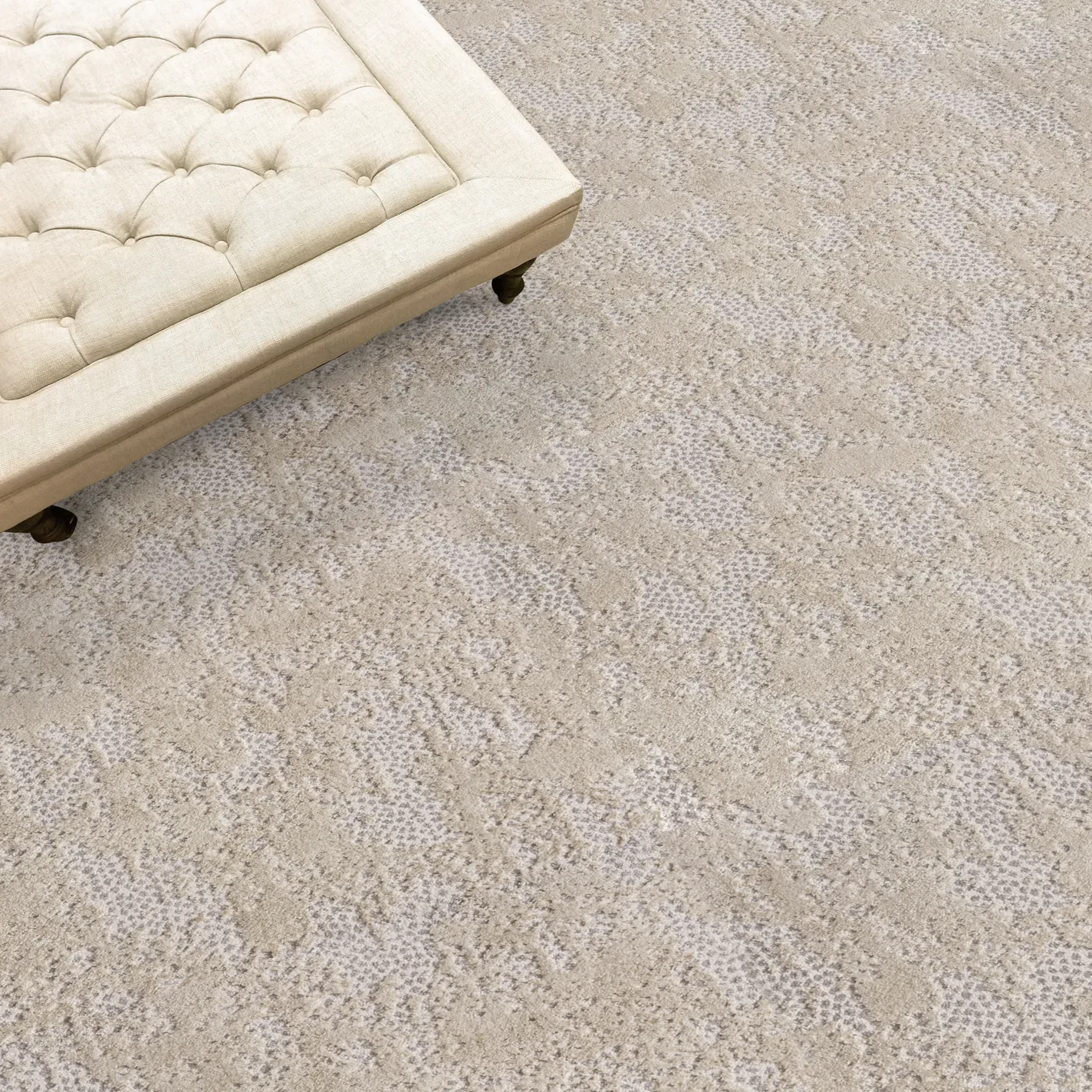 A cream tufted ottoman with dark legs rests on a light beige and grey patterned rug with an abstract, mottled design.