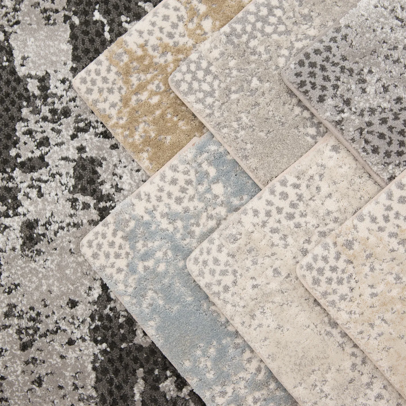 Overhead view of six rectangular carpet samples fanned out diagonally over a larger abstract grey rug. The samples feature speckled patterns in shades of beige, light grey, dark grey, light blue, and cream.