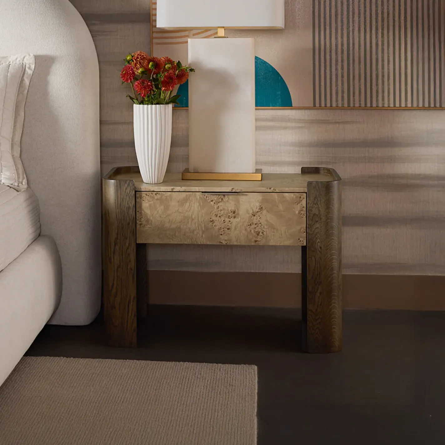A modern bedroom nightstand made of dark wood and burl wood, topped with a white fluted vase of red-orange flowers and a rectangular lamp. An upholstered headboard and abstract art are in the background.