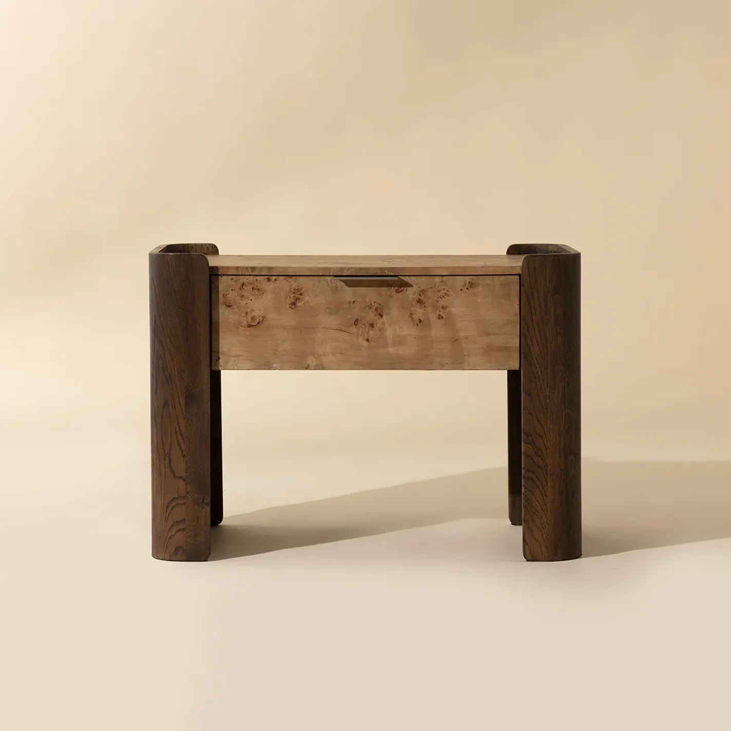 A modern wooden nightstand with a light burl wood drawer and top, framed by dark, curved wooden legs, against a light beige background.