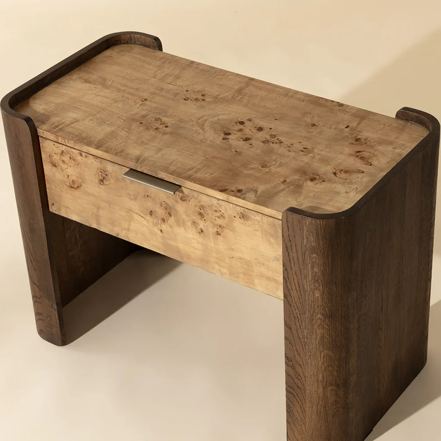 A modern two-tone wooden side table with dark brown frame and legs, and a lighter burl wood top and drawer with a recessed silver handle.