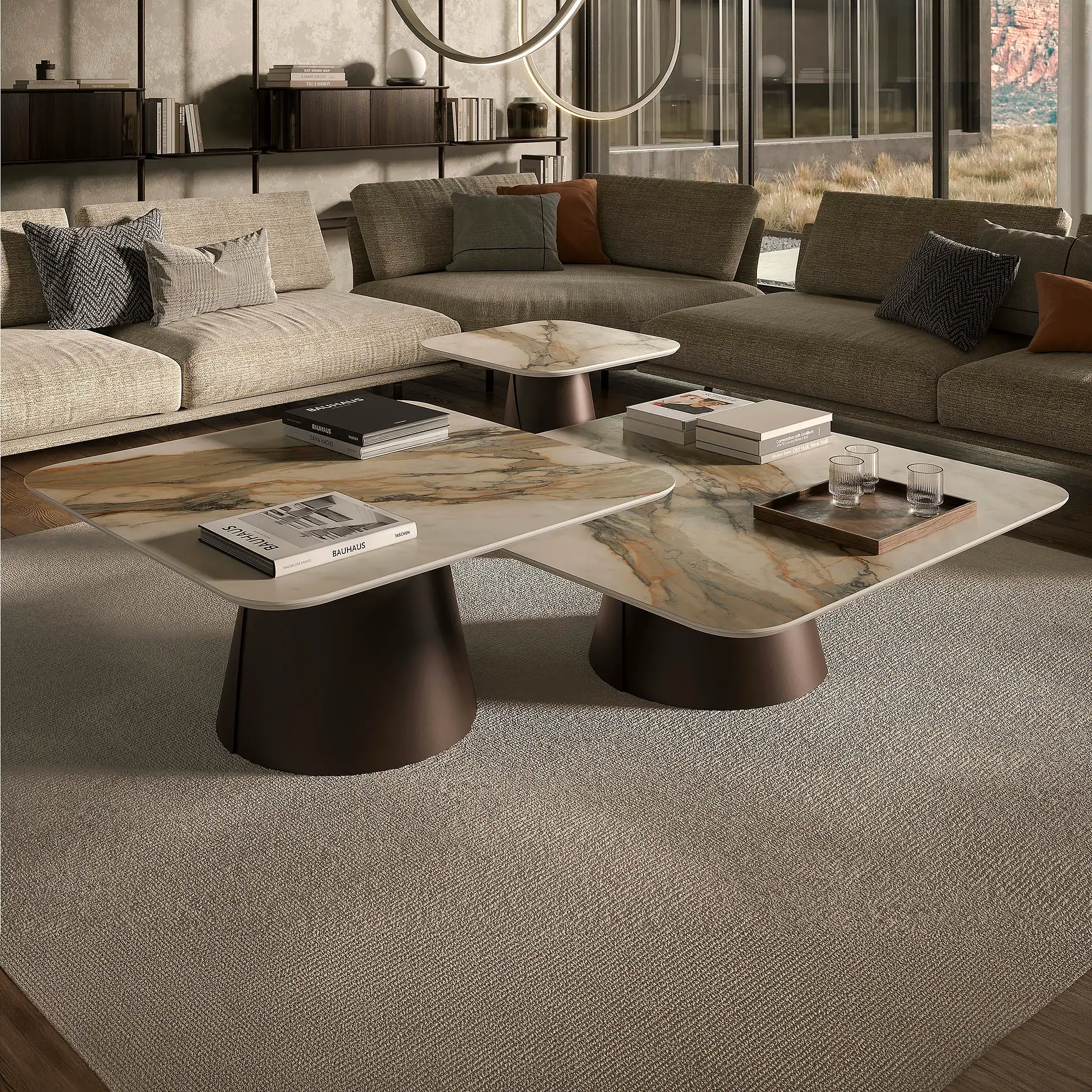 A modern living room with a large L-shaped sectional sofa, marble-topped coffee tables, and a textured rug. Books and glasses are on the tables, and a shelving unit is in the background. Large windows overlook a dry, grassy landscape.