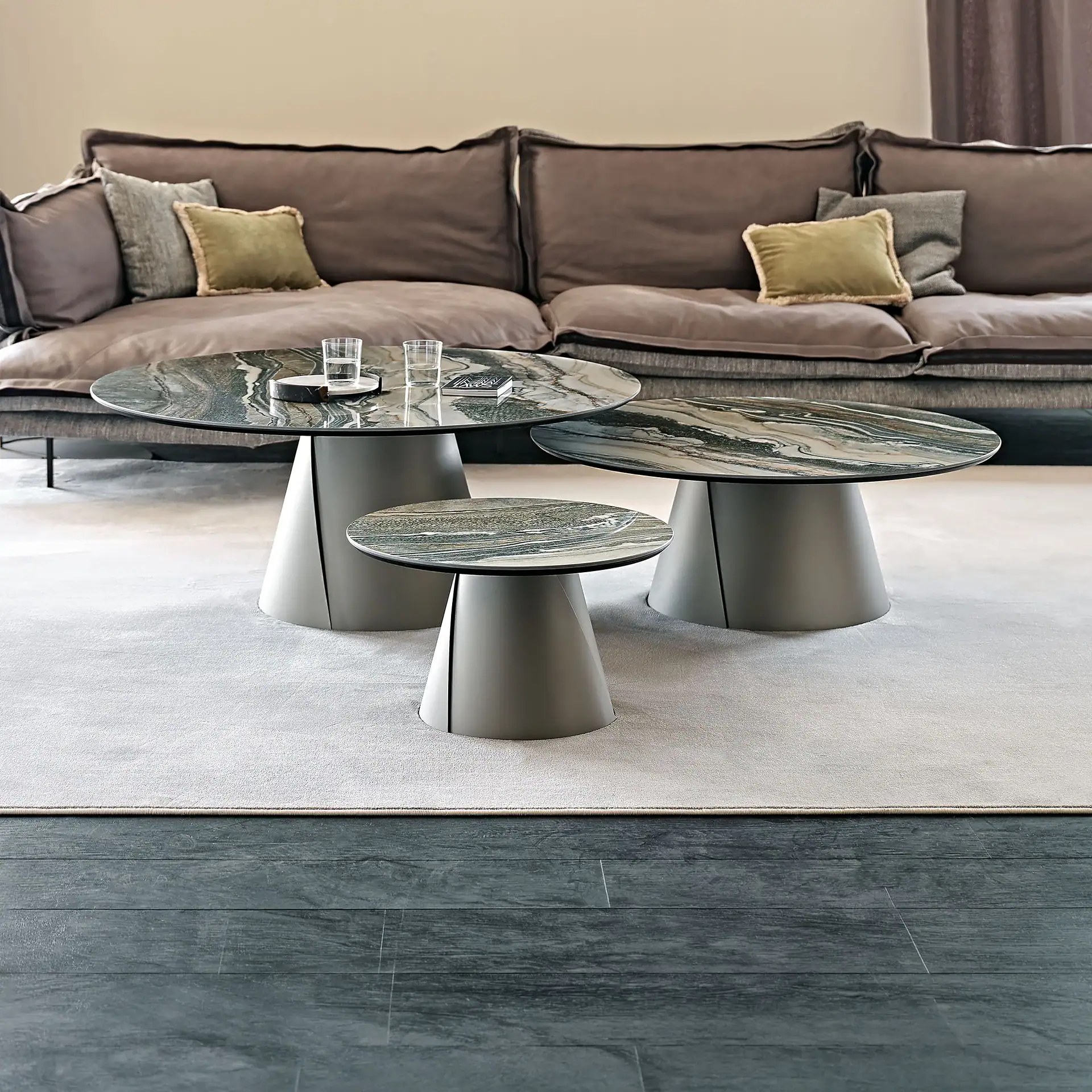 Three modern nesting coffee tables with marble-patterned tops and gray conical bases, placed on a light gray rug in front of a dark gray sofa.
