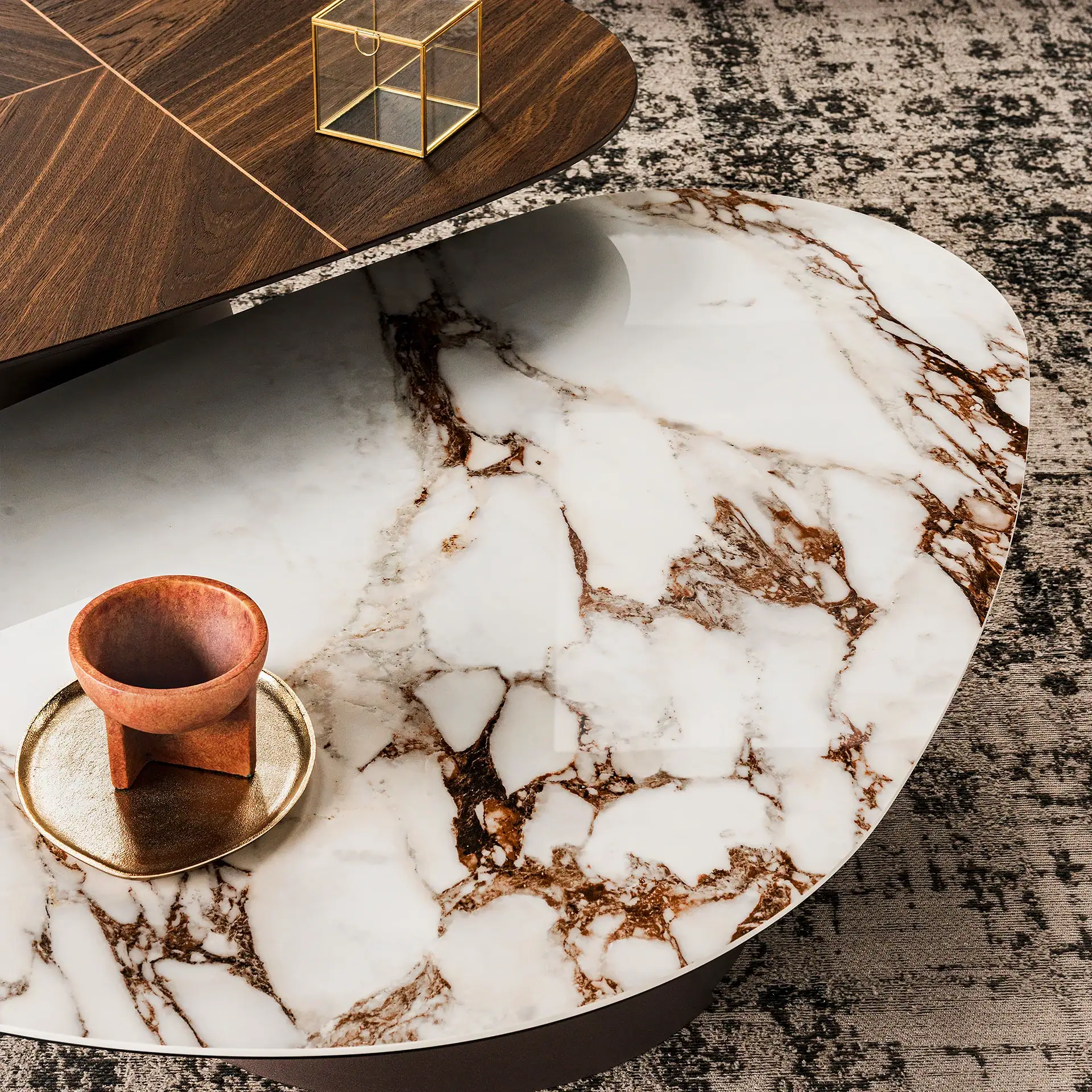 Two overlapping, kidney-shaped coffee tables on a patterned rug. One table is dark wood with brass inlay, holding a golden glass box. The other is white marble with brown veining, holding a terracotta bowl on a golden tray.