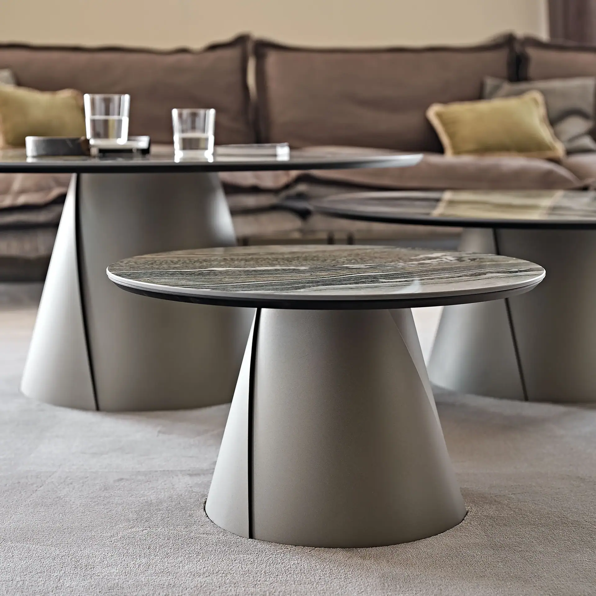 Two modern round coffee tables with marble-patterned tops and conical bases on a light grey carpet, with a brown sofa and pillows in the background.