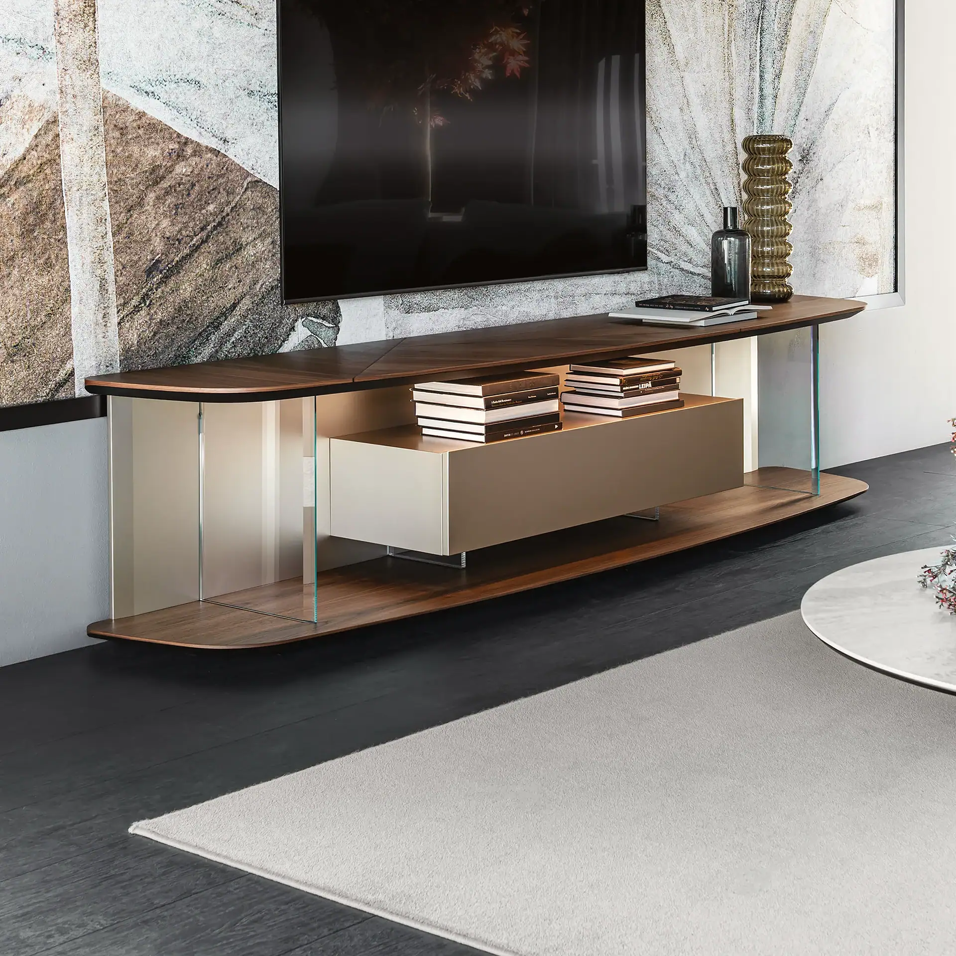 A modern media console with dark wood shelves, clear glass supports, and a light-colored floating cabinet. A flat-screen TV is mounted above it, and decorative items and books are displayed on the shelves.