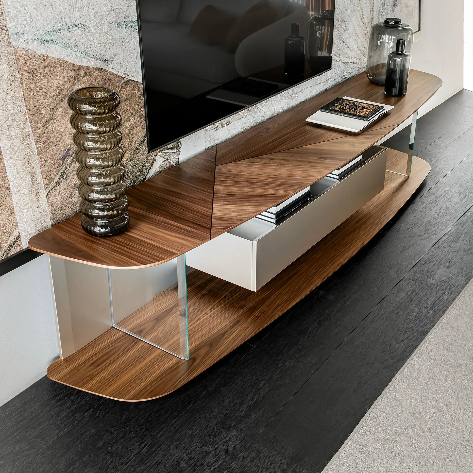 A modern walnut wood media console with glass supports, a central drawer, and decorative glass vases, positioned below a wall-mounted TV on a textured wall.