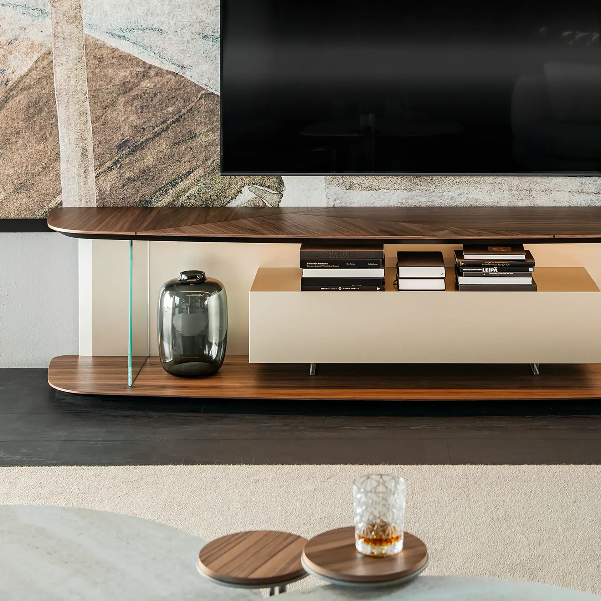 A modern living room with a large TV, a sleek dark wood and cream media console, abstract wallpaper, and a glass of whiskey on a coffee table.