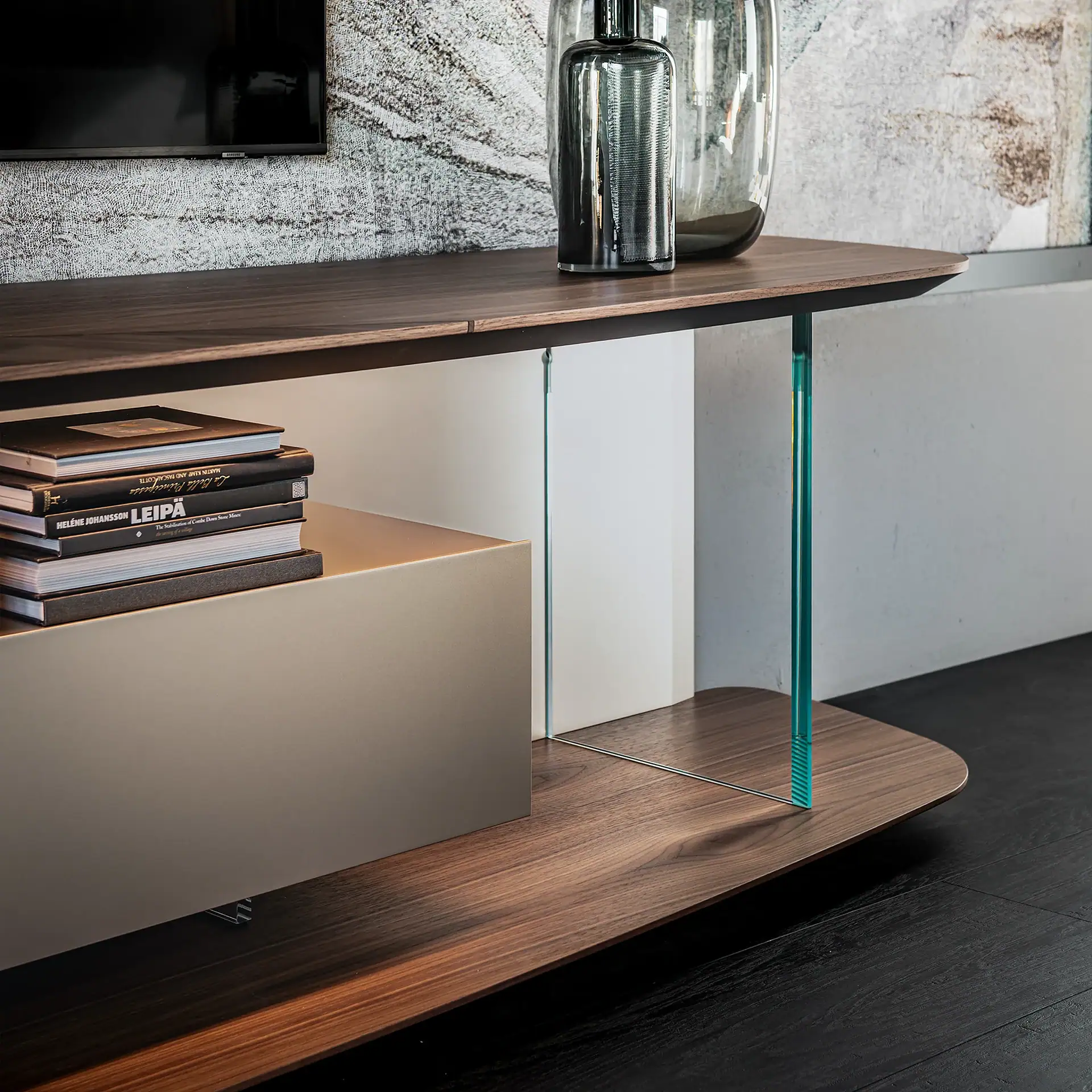 A modern media console with dark wood shelves, clear and blue-green glass supports, and a light beige drawer. Two glass vases and a stack of books are on the shelves.