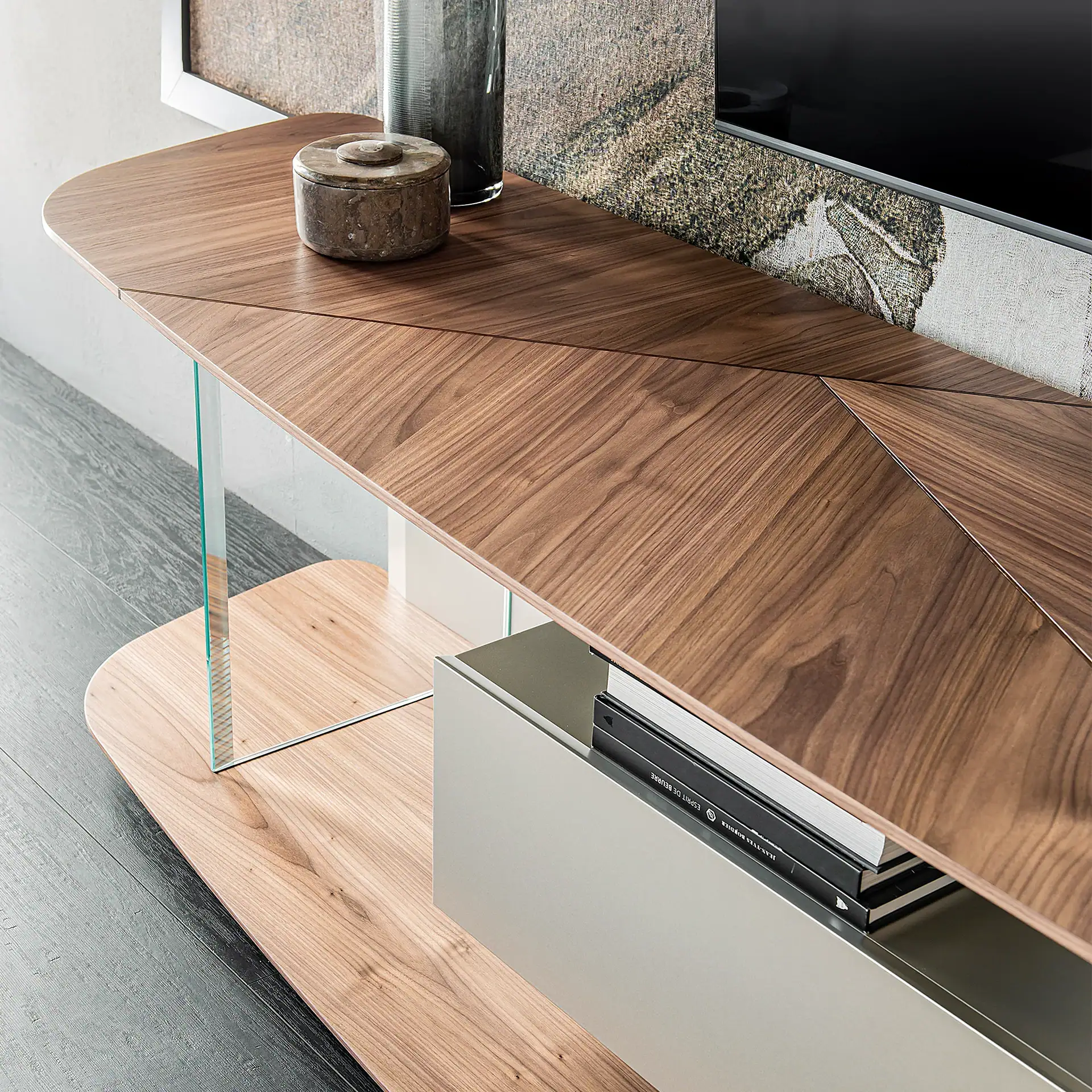 A modern media console with a geometric walnut top, glass supports, and a light wood base, featuring a partially open drawer with books.