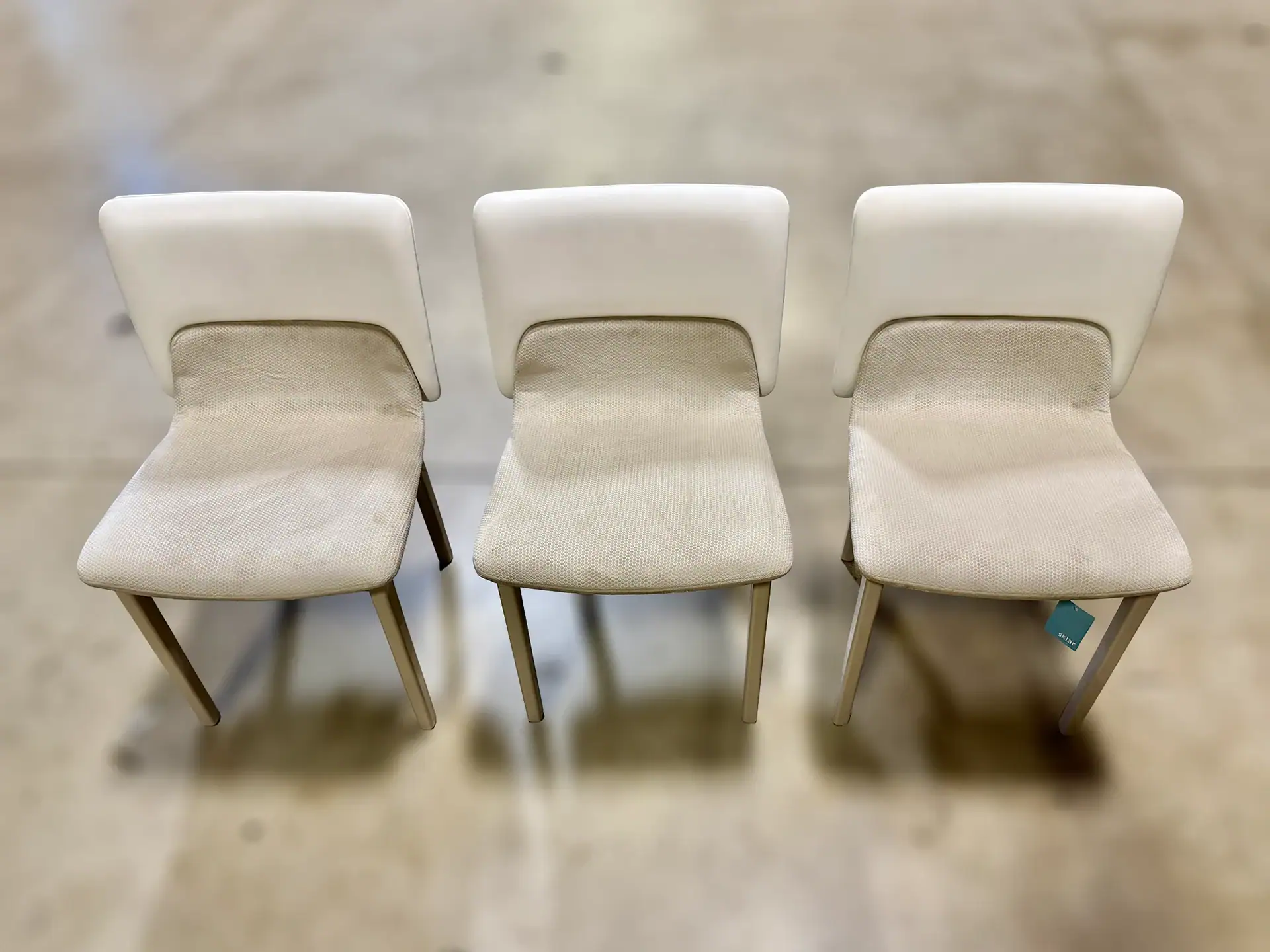 Three white Nora dining chairs with wooden legs on a concrete floor.