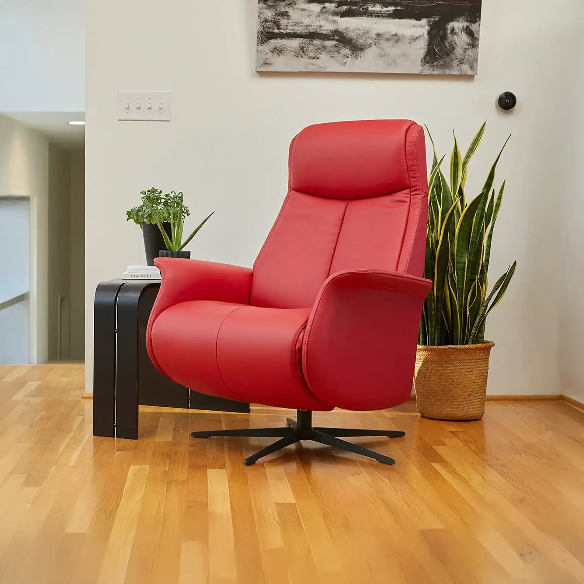 A bright red modern armchair with a black base on a hardwood floor, flanked by a black side table with plants and a large snake plant in a woven basket. A white wall with light switches, abstract art, and a smart thermostat is in the background.