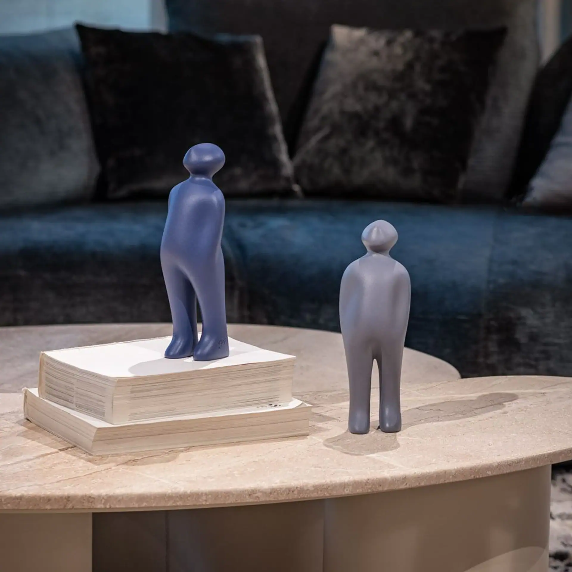 Two abstract human-like figurines, one blue-purple and one light gray, stand on a marble coffee table with books. A dark sofa is in the blurred background.