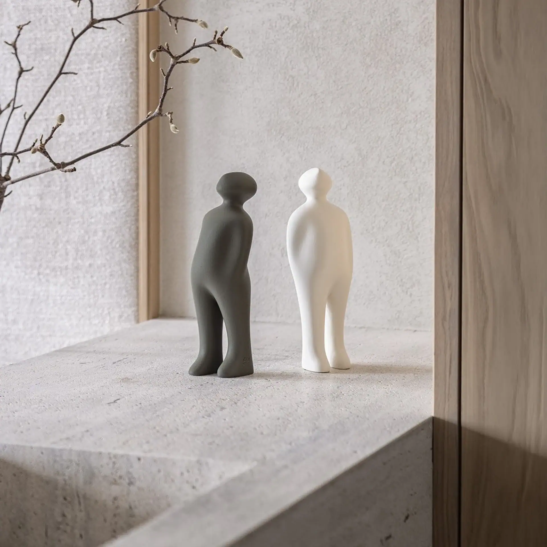 Two abstract, human-like figures, one dark gray and one off-white, standing on a textured surface next to a branch with buds.